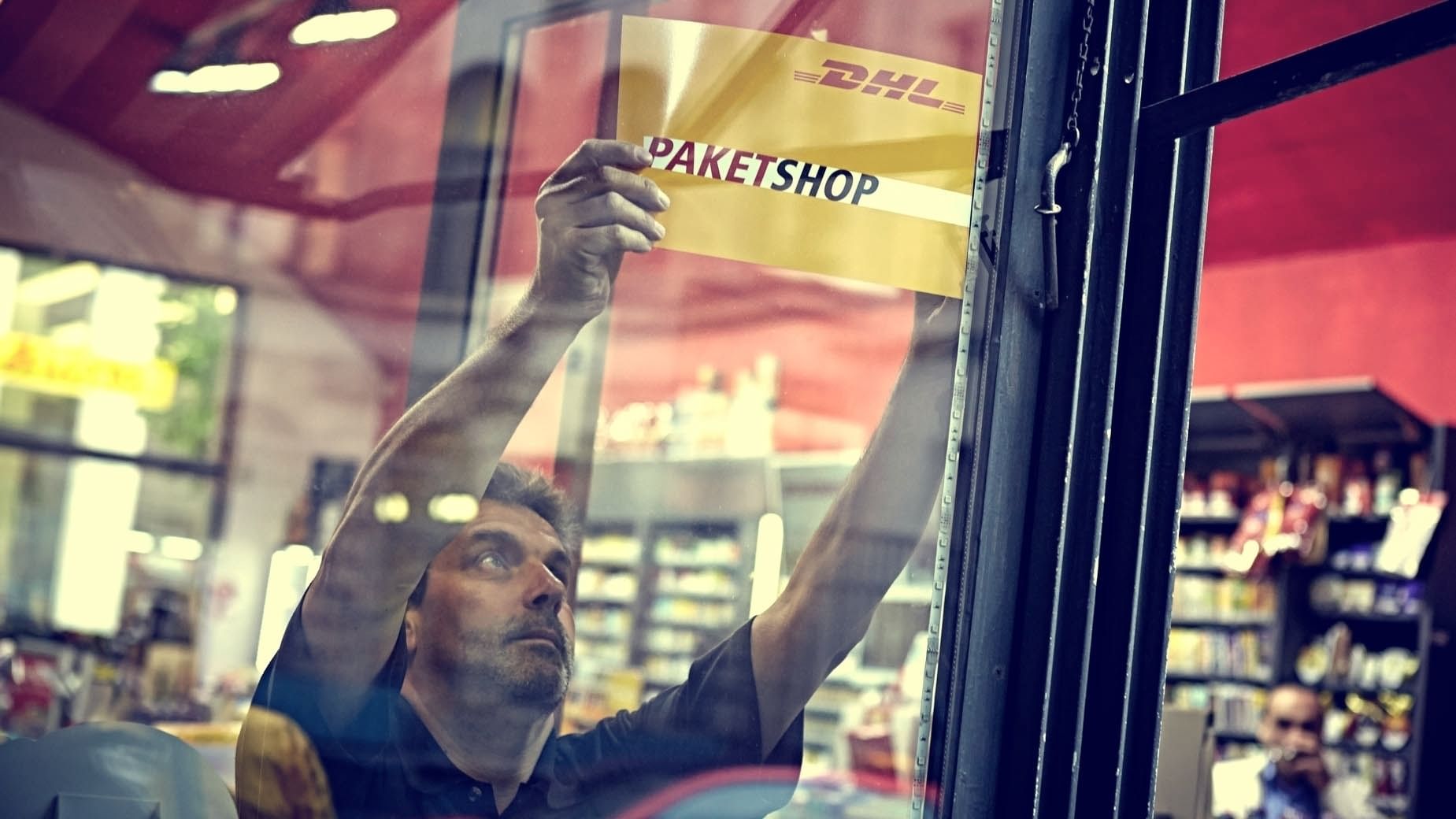 Ein Mann klebt ein Paketshop-Schild der DHL in das Schaufenster eines Ladens.