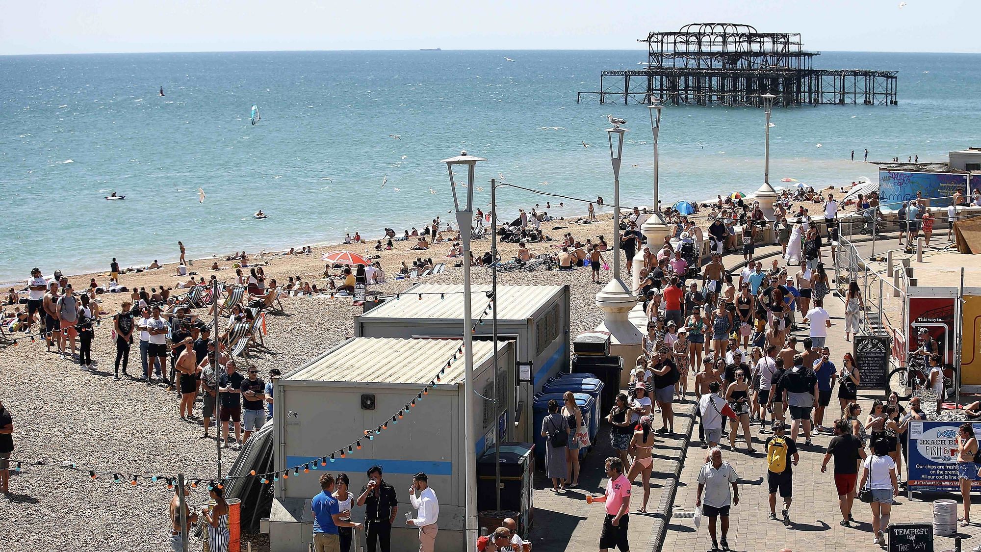 Am Strand in Brighton stehen eine Menge Menschen in einer Schlange vor einem Kiosk, andere laufen eng nebeneinander über die Promenade. Mundschutz trägt dabei niemand, der oder die zu sehen ist. 