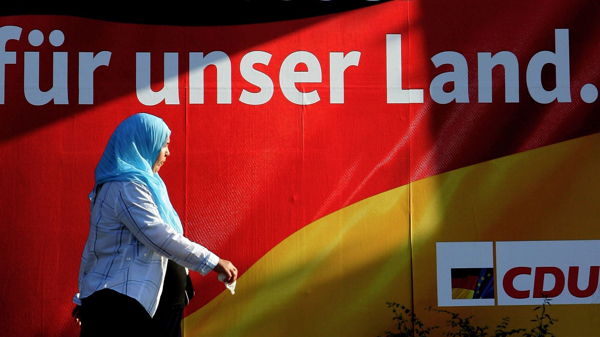 Wir sehen eine Frau mit Kopftuch, die vor einer schwarz rot goldenen CDU-Werbung läuft, auf der steht „FÜR UNSER LAND - CDU“. 