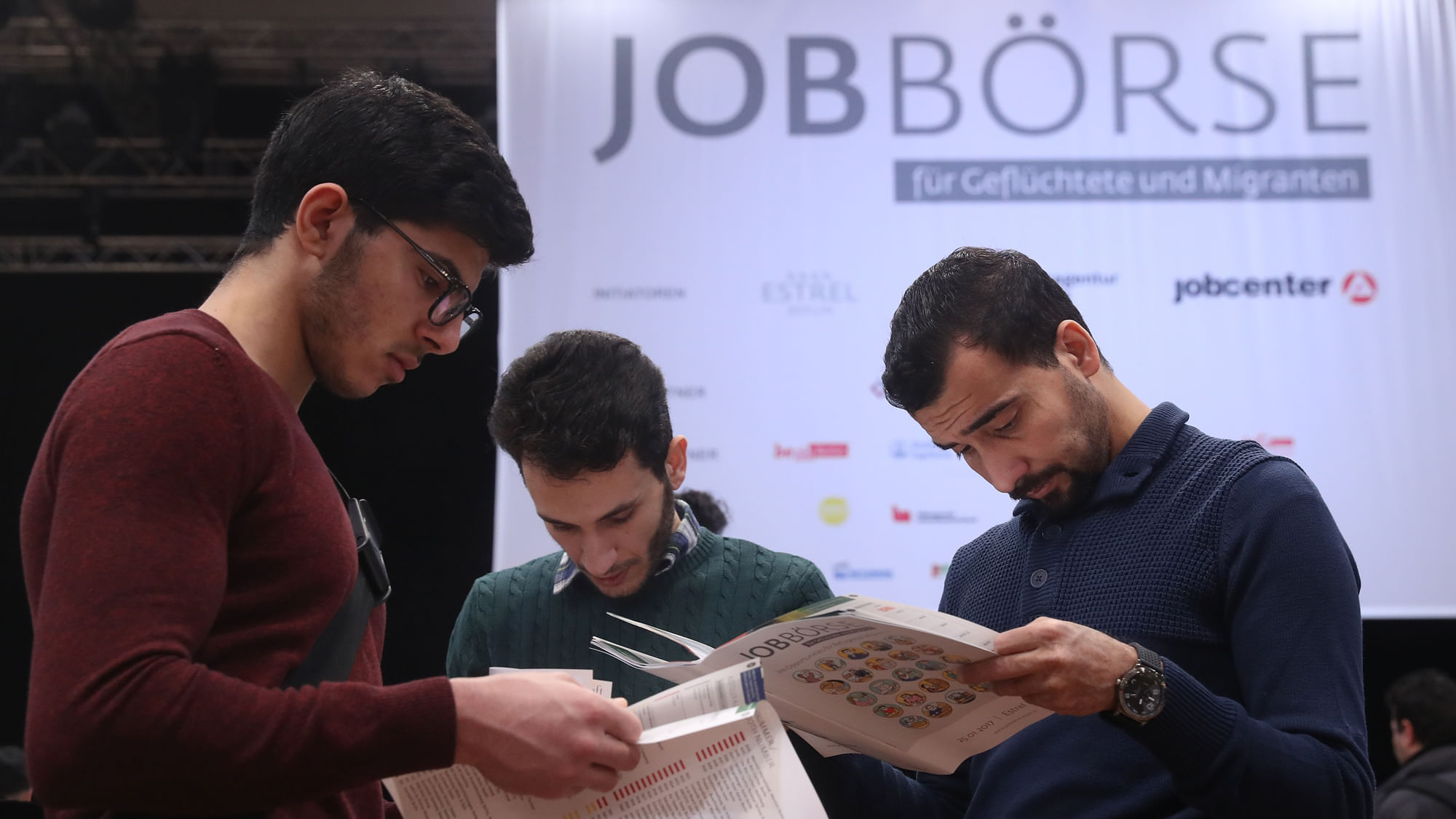 Wir sehen drei Menschen, die sich Broschüren ansehen auf einer Messe, im Hintergrund ist ein Plakat zu sehen mit der Aufschrift: JOBBÖRSE für Geflüchtete und Migranten