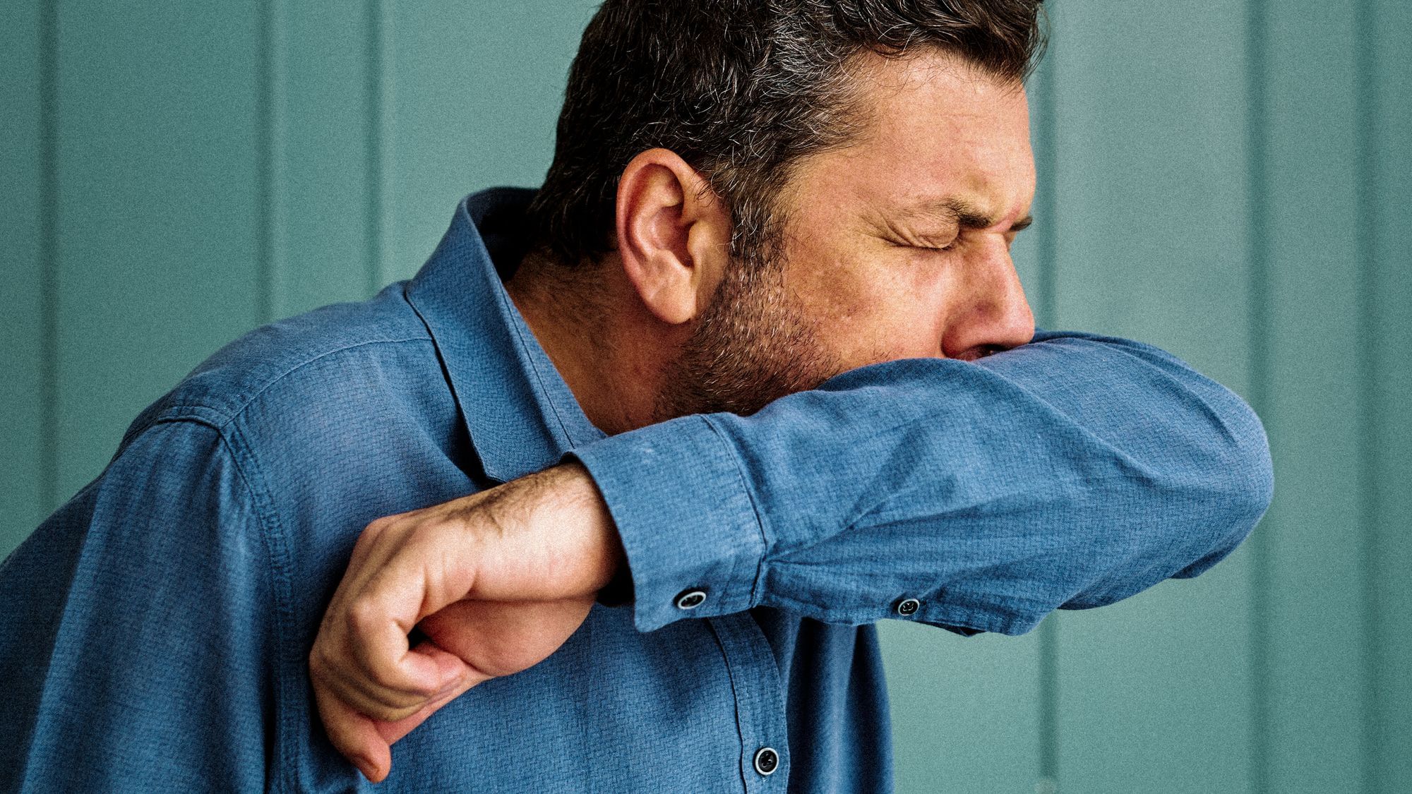 Wir sehen eine männlich gelesene Person, die sich in die Armbeuge hustet. 