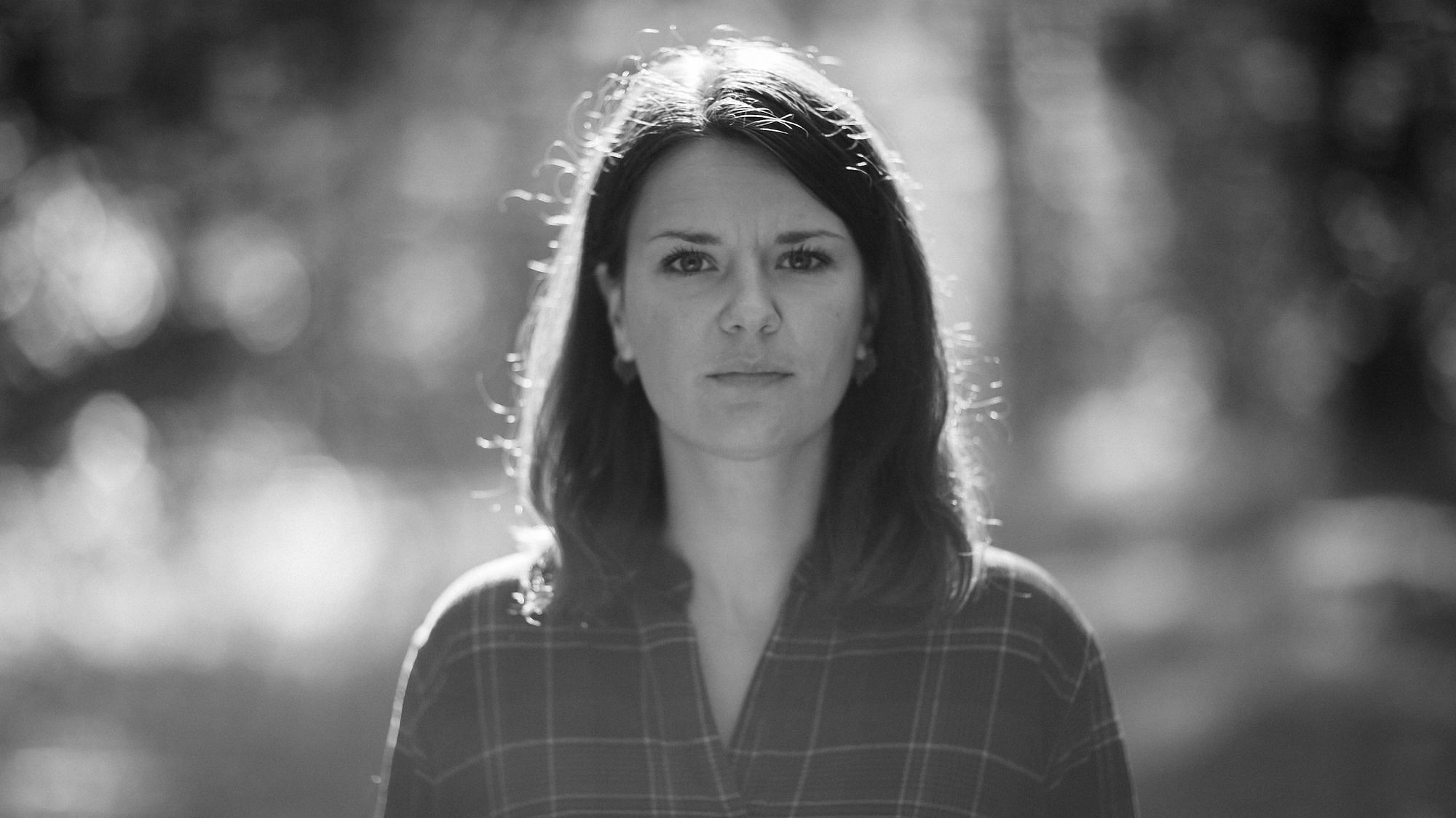 Wir sehen die Autorin und sie schaut im Gegenlicht und umgeben von Wald in die Kamera.