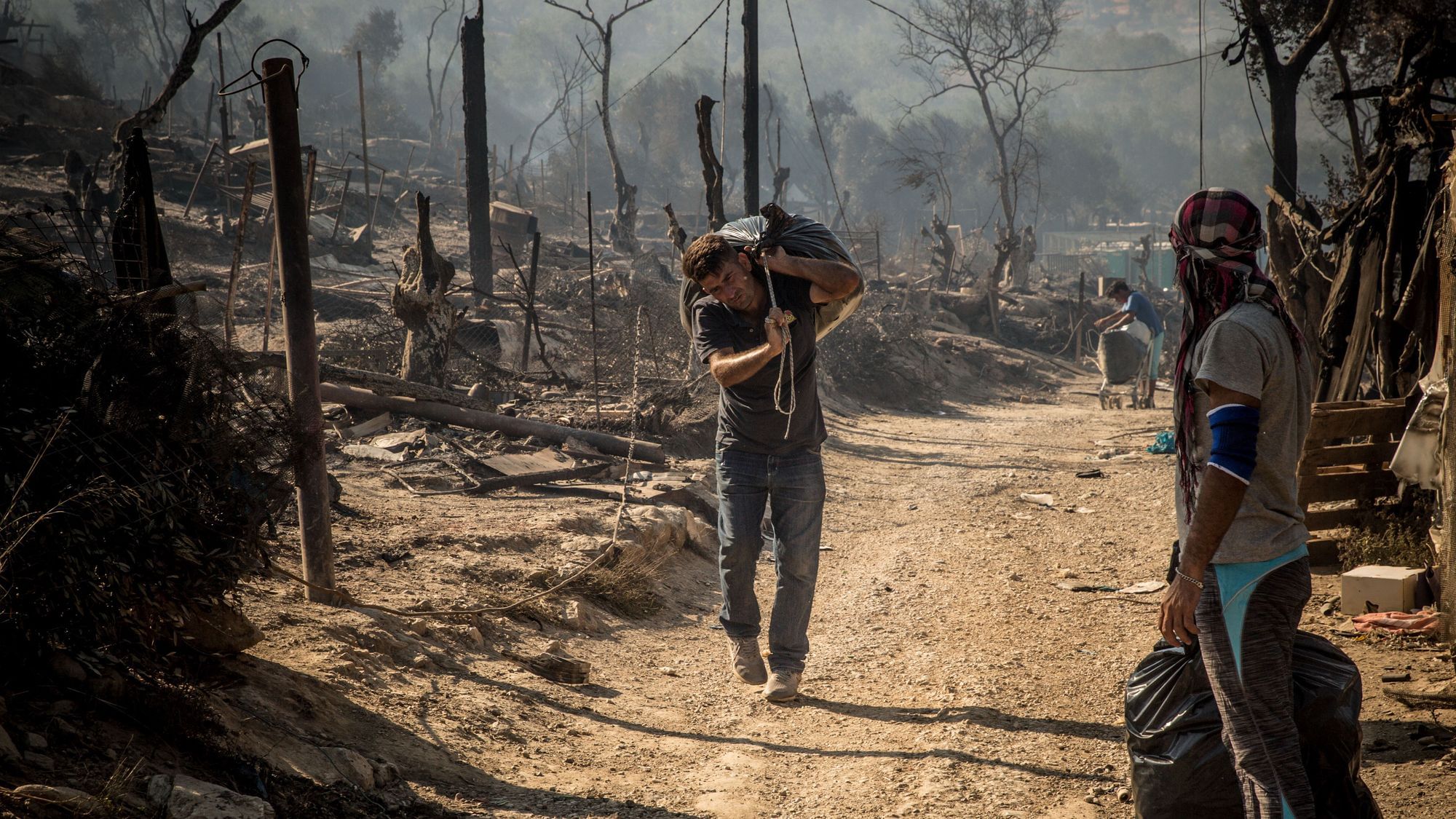 Wir sehen eine Person, die durch ein abgebranntes Lager läuft und auf dem Rücken einen großen Plastiksack trägt – vermutlich das letzte Hab und Gut. 