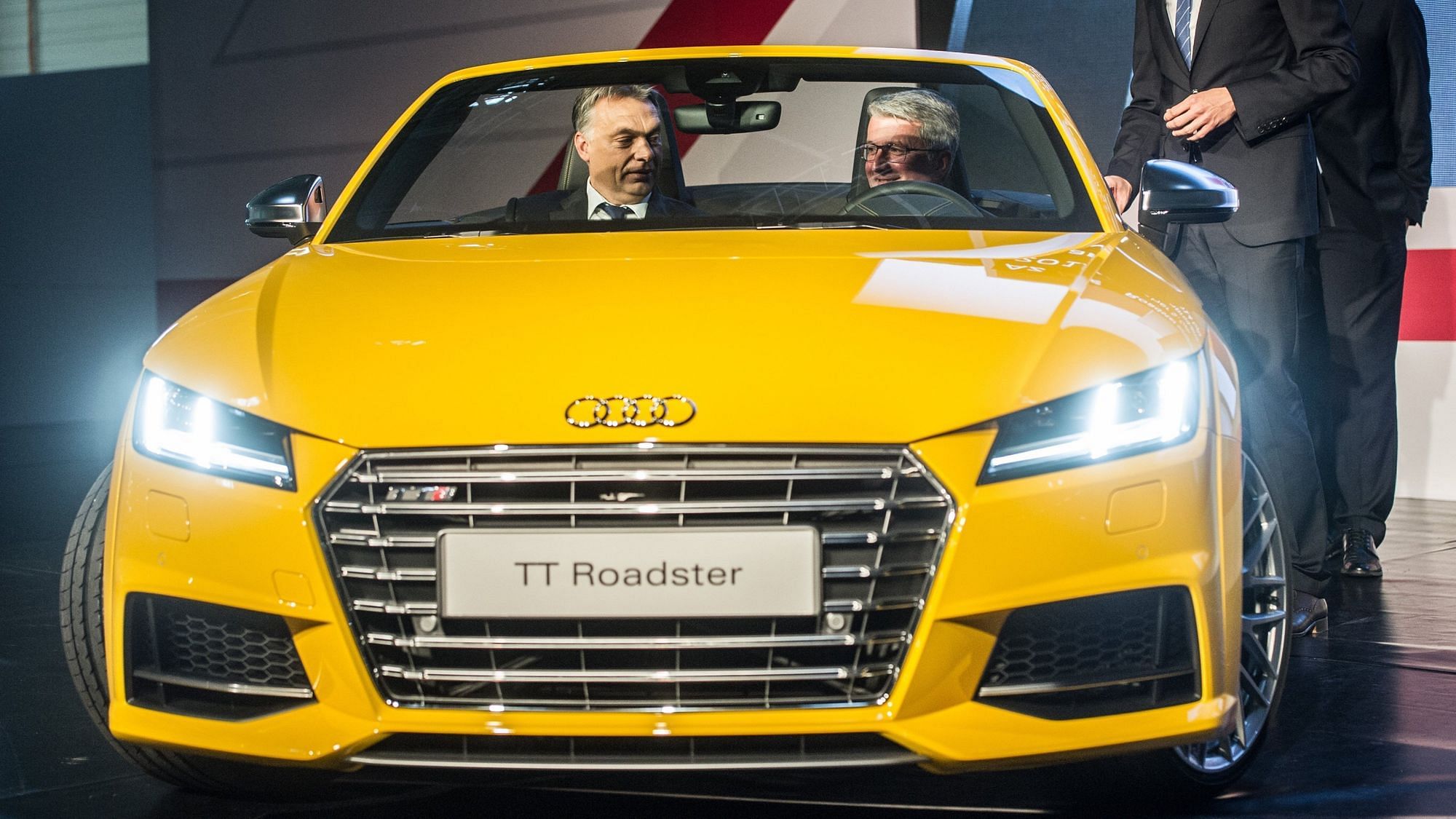 Wir sehen Orban mit einem zweiten Herrn in einem gelben Audi TT Roadster sitzen. 