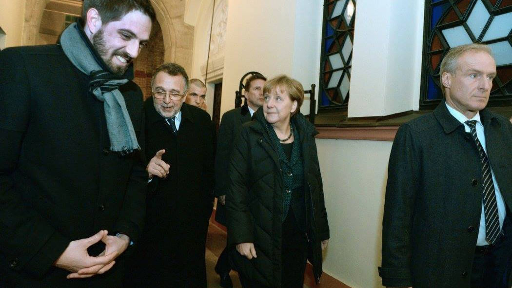 Wir sehen Angela Merkel umgeben von mehreren Männern im Anzug durch eine Synagoge laufen. 