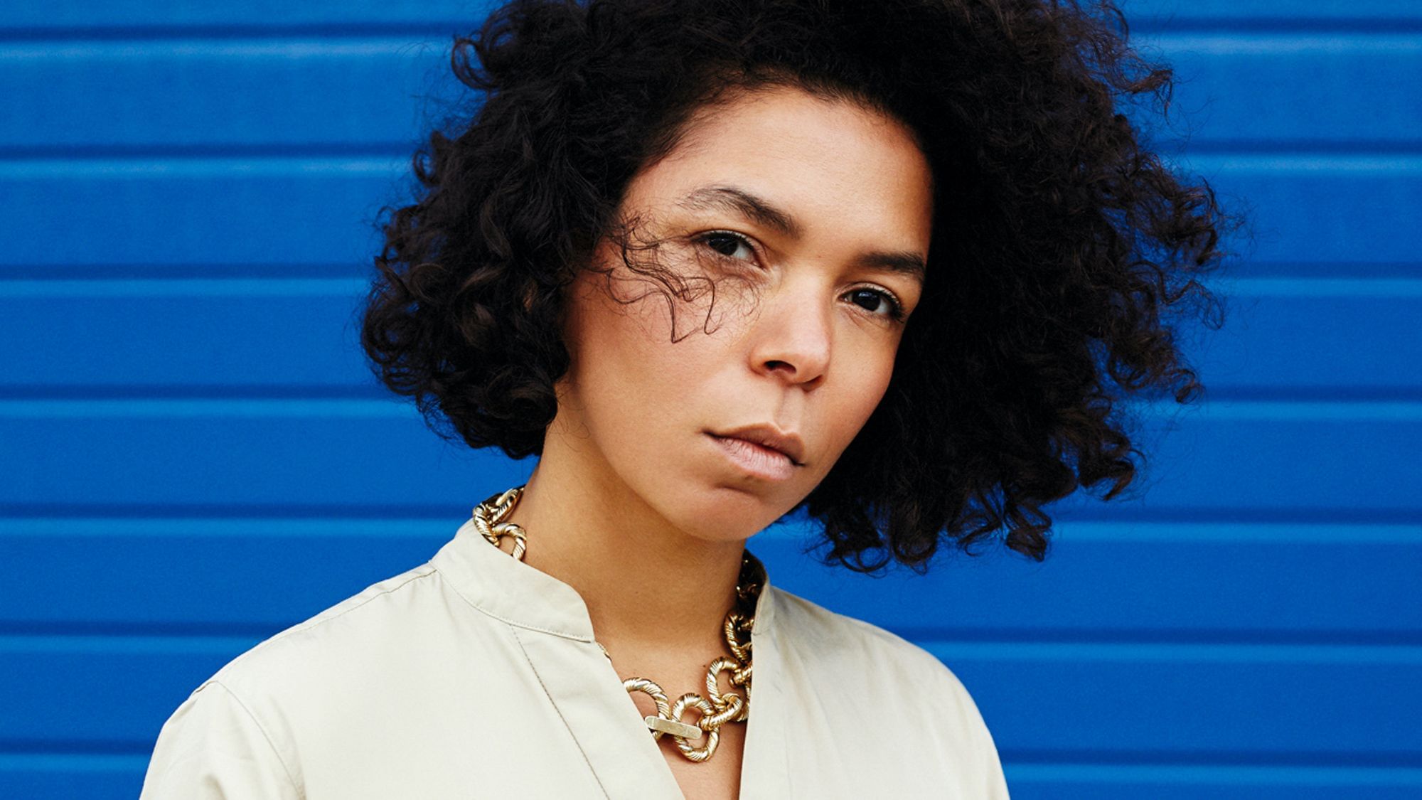 Wir sehen die Autorin Katharina Warda, wie sie heute aussieht. Locken, braune Augen und einen relativ ersten Gesichtsausdruck, fotografiert vor einer blauen Wand.