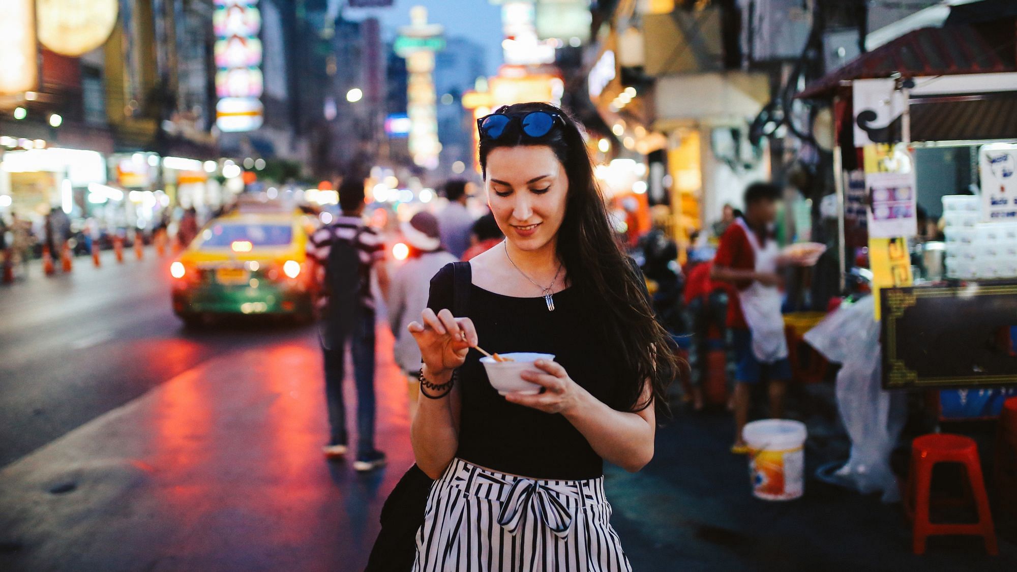 Wir sehen eine Frau, die genüßlich ein auf der Straße gekauftes Essen verspeißt, im Hintergrund Großstadtleben.