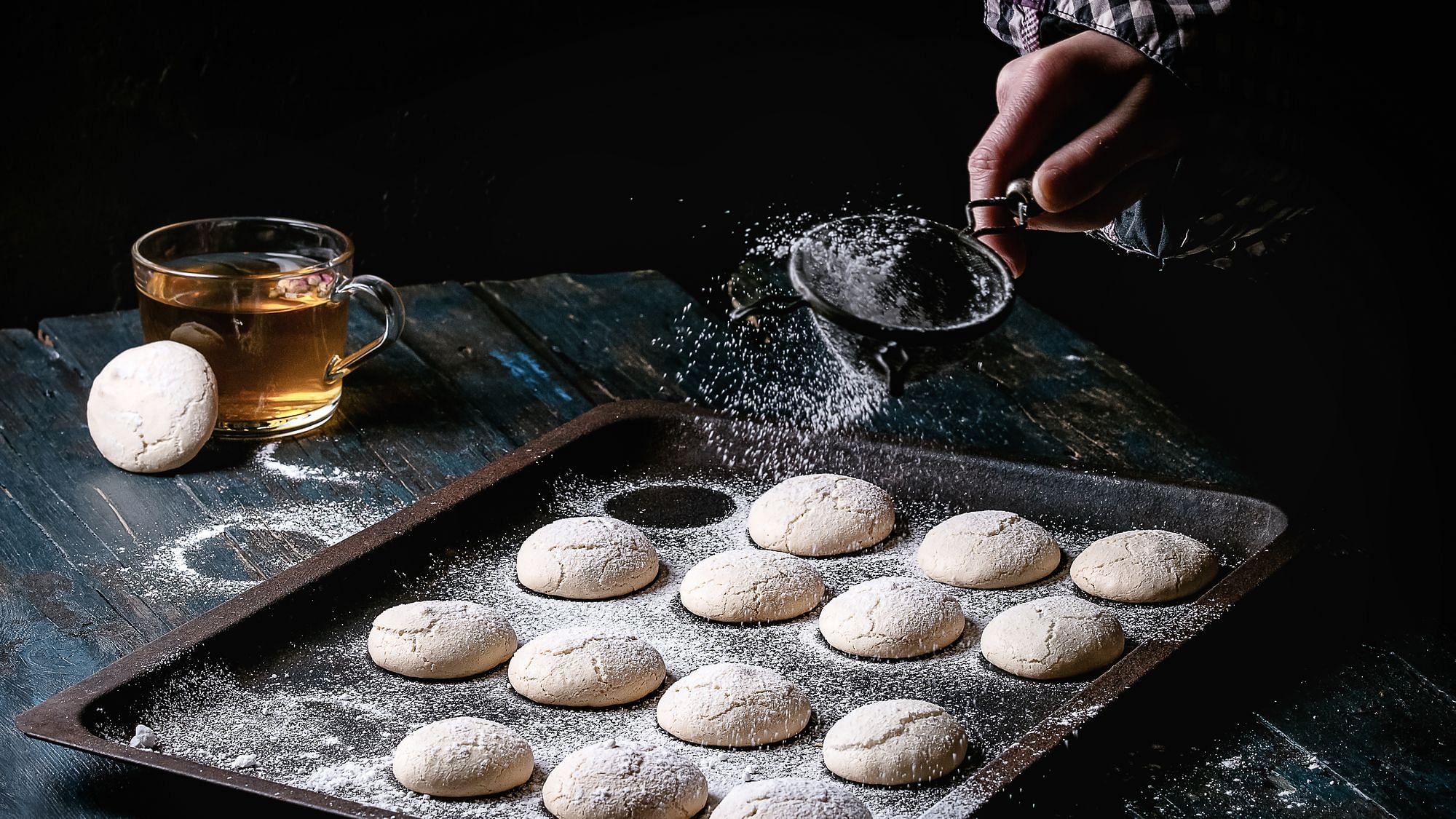 Ein Kuchenblech mit Mandelkeksen wird mit Puderzucker gepudert. Eine Glastasse Tee steht daneben.