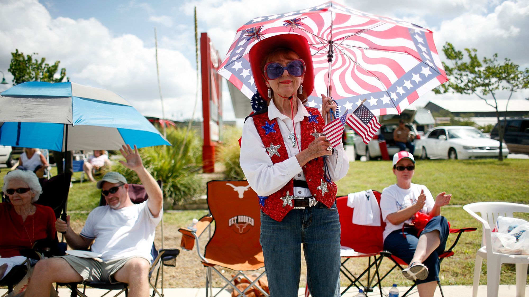 Eine Frau in roter Plüschweste mit Blauen und Silbernen Sternen sowie Sonnenbrille steht unter einen Schirm im Stil und Farbschema der US-Flagge gehalten. 