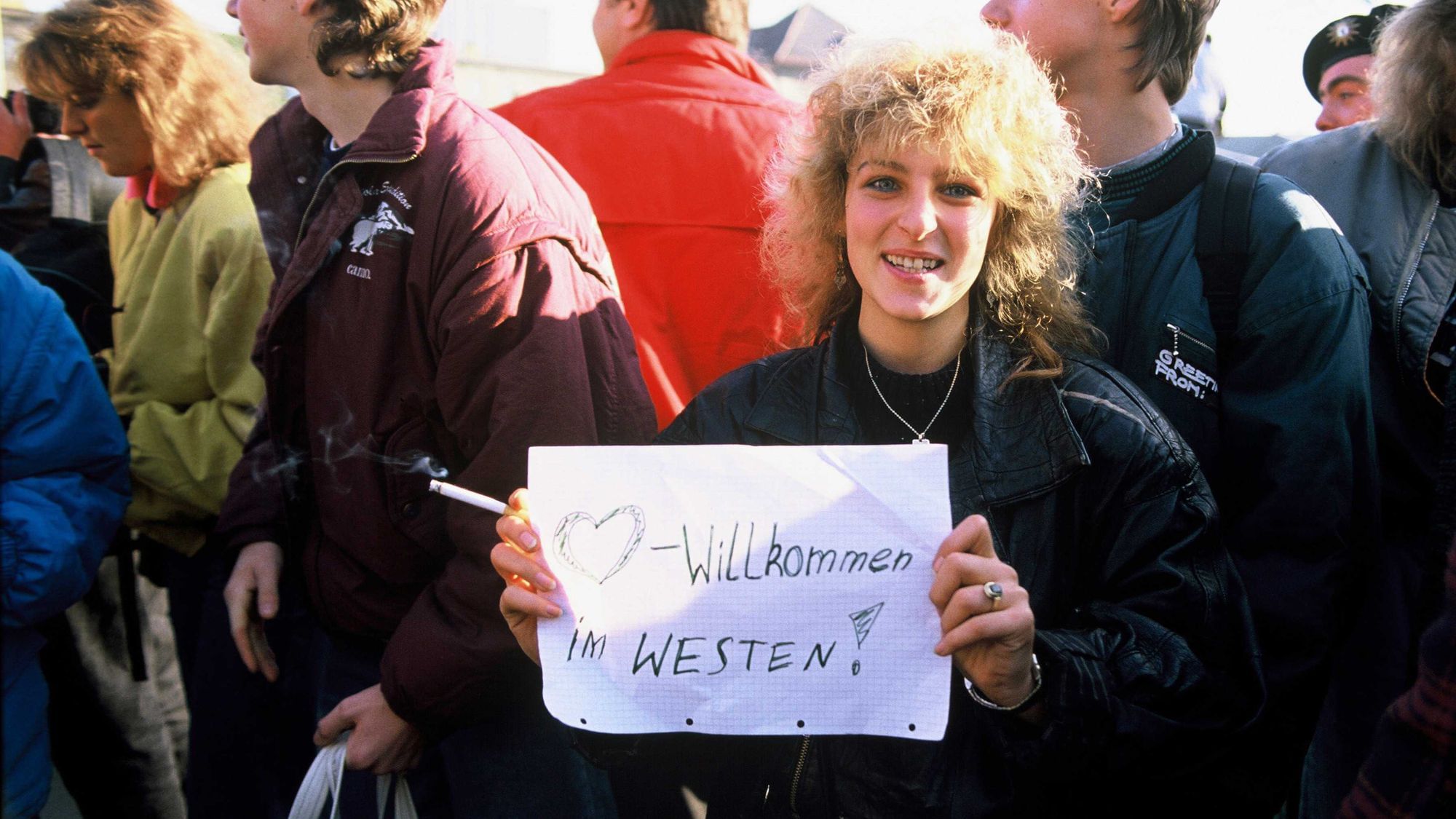 Eine Frau hält ein Blatt in den Händen auf welchem „*Herz-Zeichen* Willkommen im Westen“ steht. Das Bild is klar durch Kleidung und Haarschnitt der Frau wie auch der Menschen und Emotionen um sie herum dem Fall der Mauer zuortbar. 