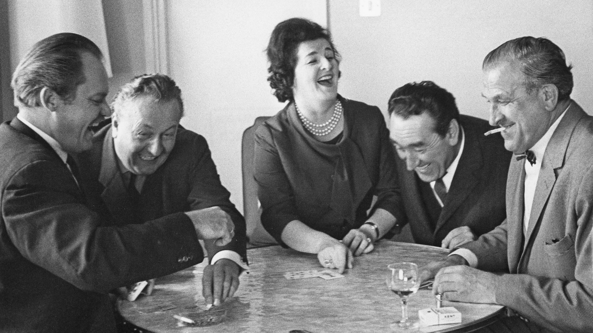 Eine Gruppe Männer sitzt mit einer Frau zusammen am Tisch, sie rauchen, trinken und lachen sich einen Ast ab.