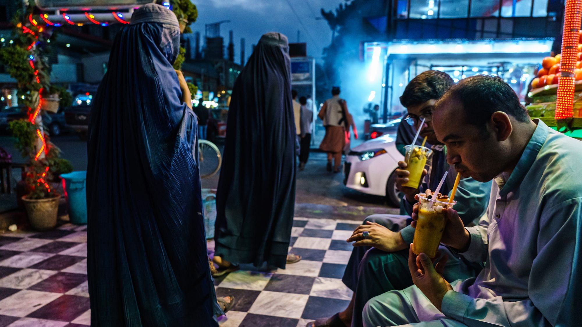 Abendliche Alltagsszene, zwei Frauen im Burqa laufen an zwei Männern vorbei welche Fruchtsmoothies trinken, unweit davon sehen wir im Hintergrund eine bewaffnete Person die uns den Rücken zudreht.