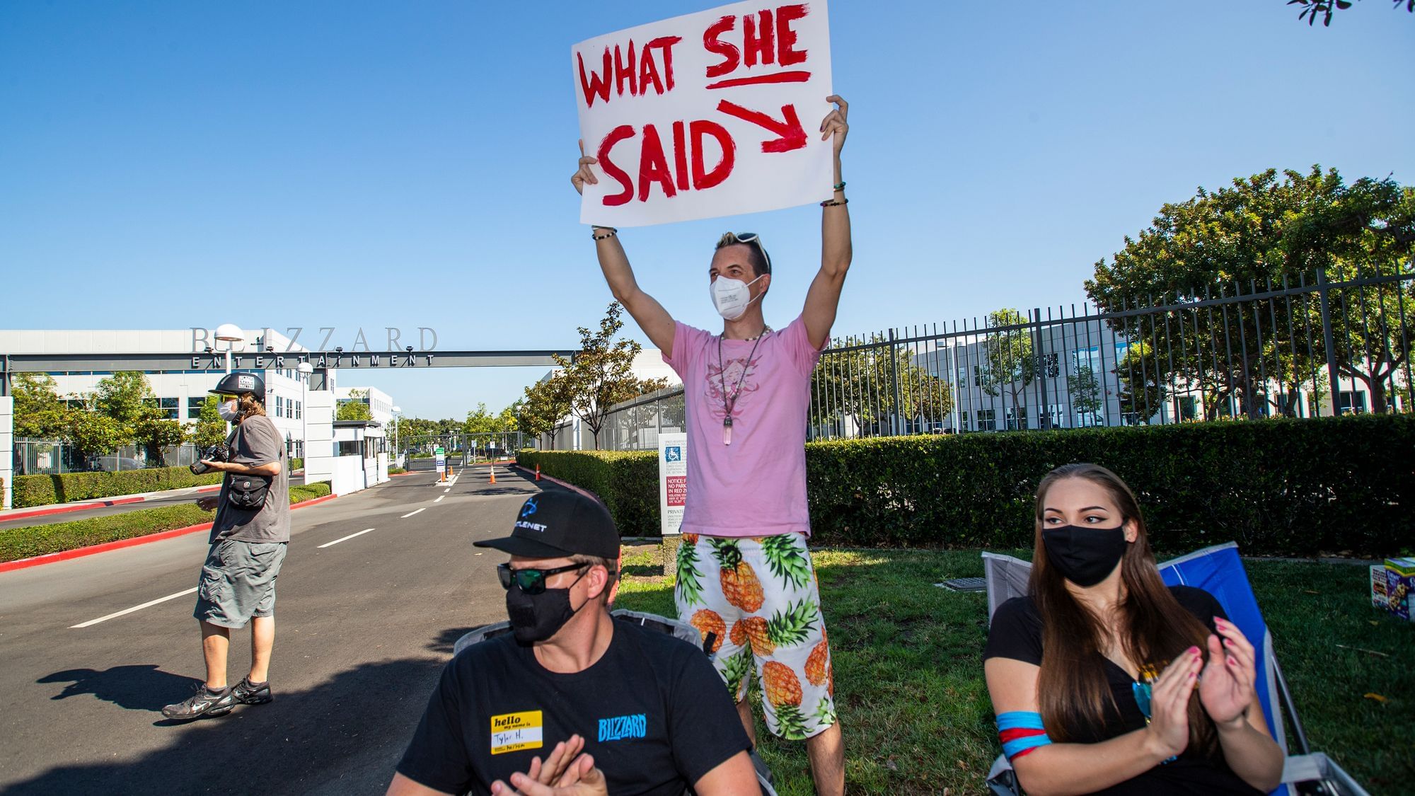 Ein Mann hält ein Schild mit der Aufschrift „What she said“ hoch, vor den Toren von Blizzard Activision. Vor ihm sitzt eine Frau und ein Mann.