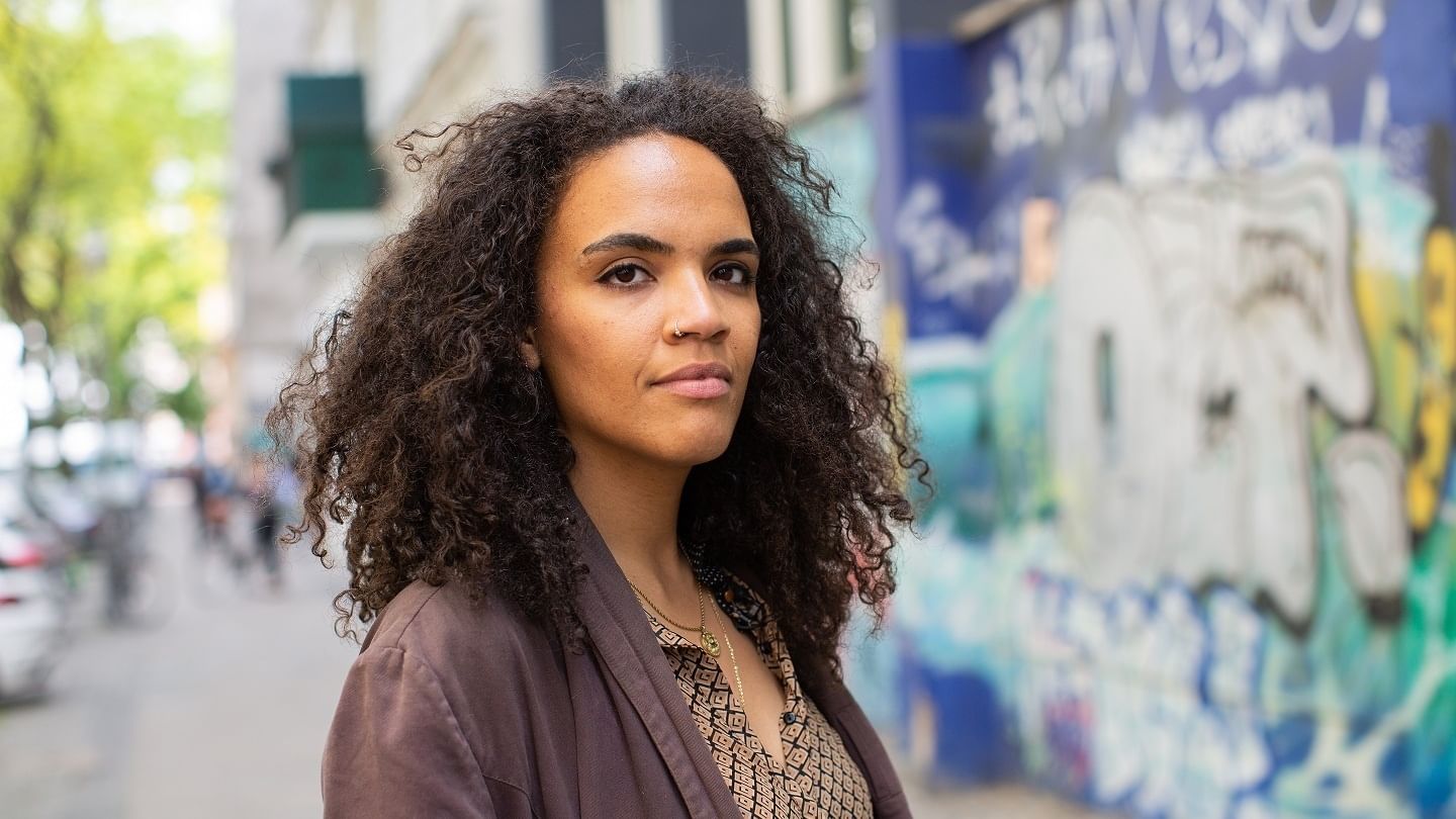 Porträt der Autorin Ciani-Sophia Hoeder, sie steht mittig auf einem Gehsteig, links ist eine Baumbeschattete Straßenszene, rechts ist eine Wand mit Graffiti.