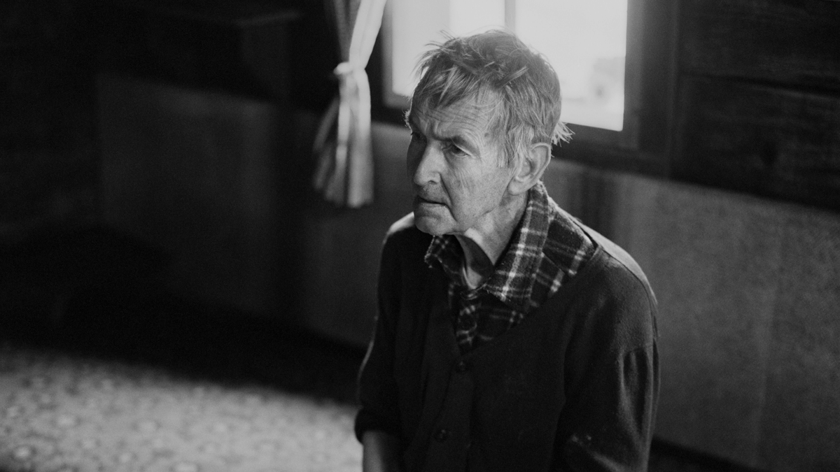 Ein Mann sitzt auf einem Bett, hinter ihm ein Fenster, duch das Licht in den dunklen Raum fällt.