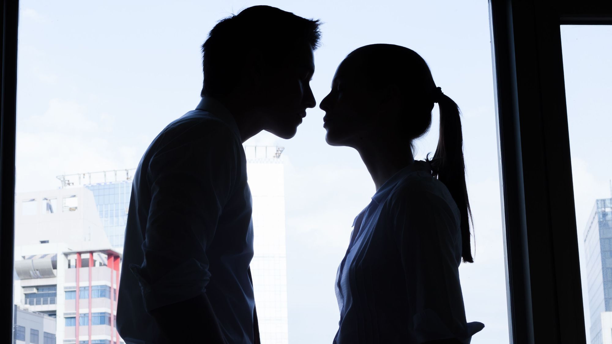 Ein Paar bestehen aus einer männlich und einer weiblich gelesenen Silhouette küsst sich. Das Fenster im Hinntergrund zeigt einen funktionalen Wolkenkratzer.
