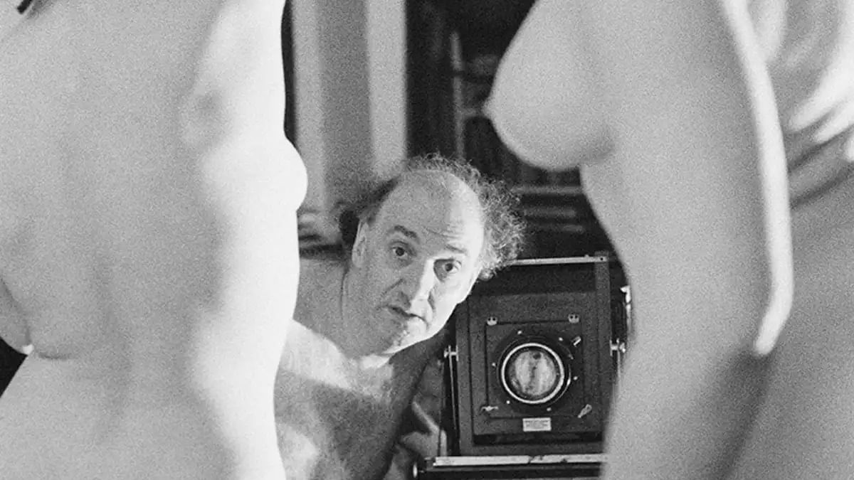 Wir blicken durch zwei nackte, weiblich gelesene Models und sehen den Fotografen der sie in Szene setzt. 