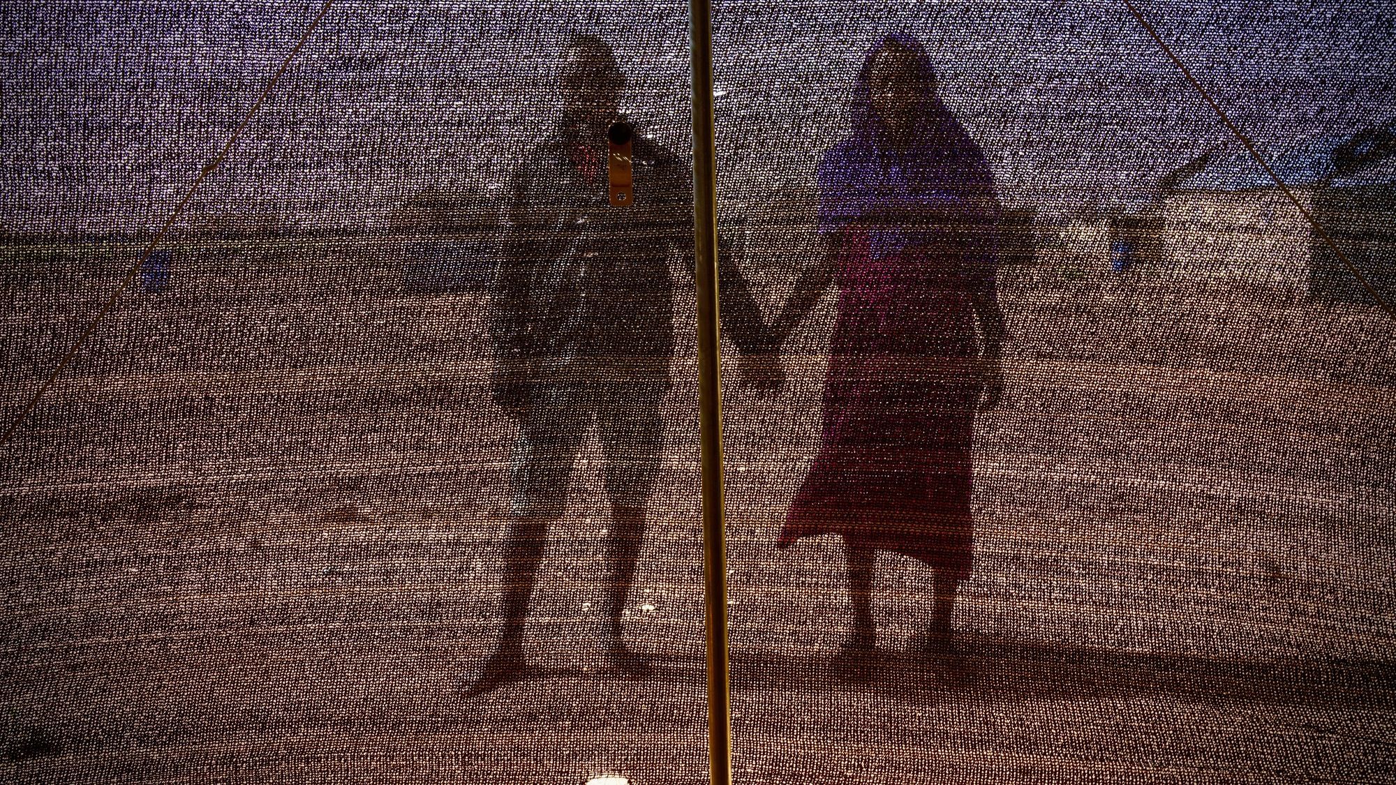 Zwei geflüchtete Menschen in Niger, scheinbar ein heterosexuelles Paar, halten Händchen, wir sehen ihre Silhouette durch ein schwarzes Maschennetz.