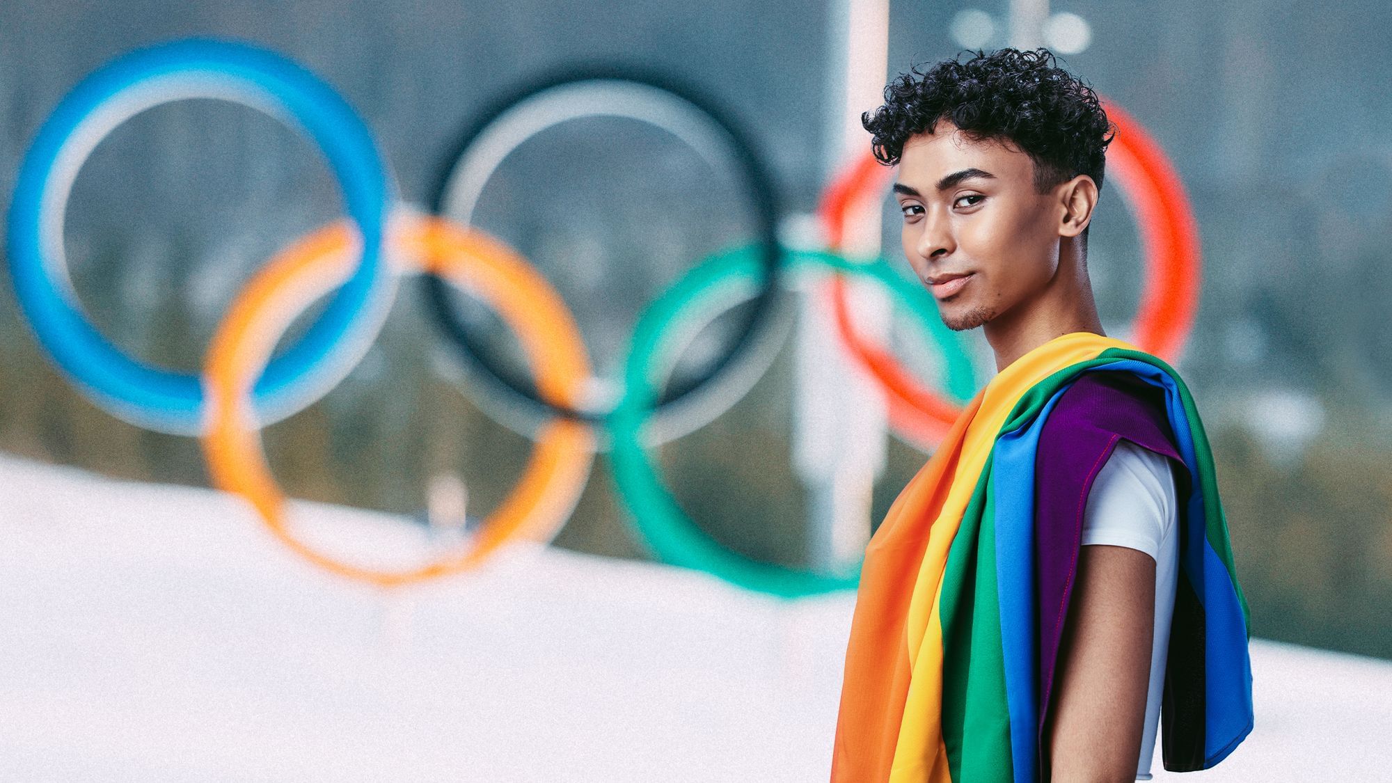 Ein junger Mann mit Regenbogenflagge über der Schulter blickt in die Kamera. Im Hintergrund sind die olympischen Ringe im Schnee zu sehen. 