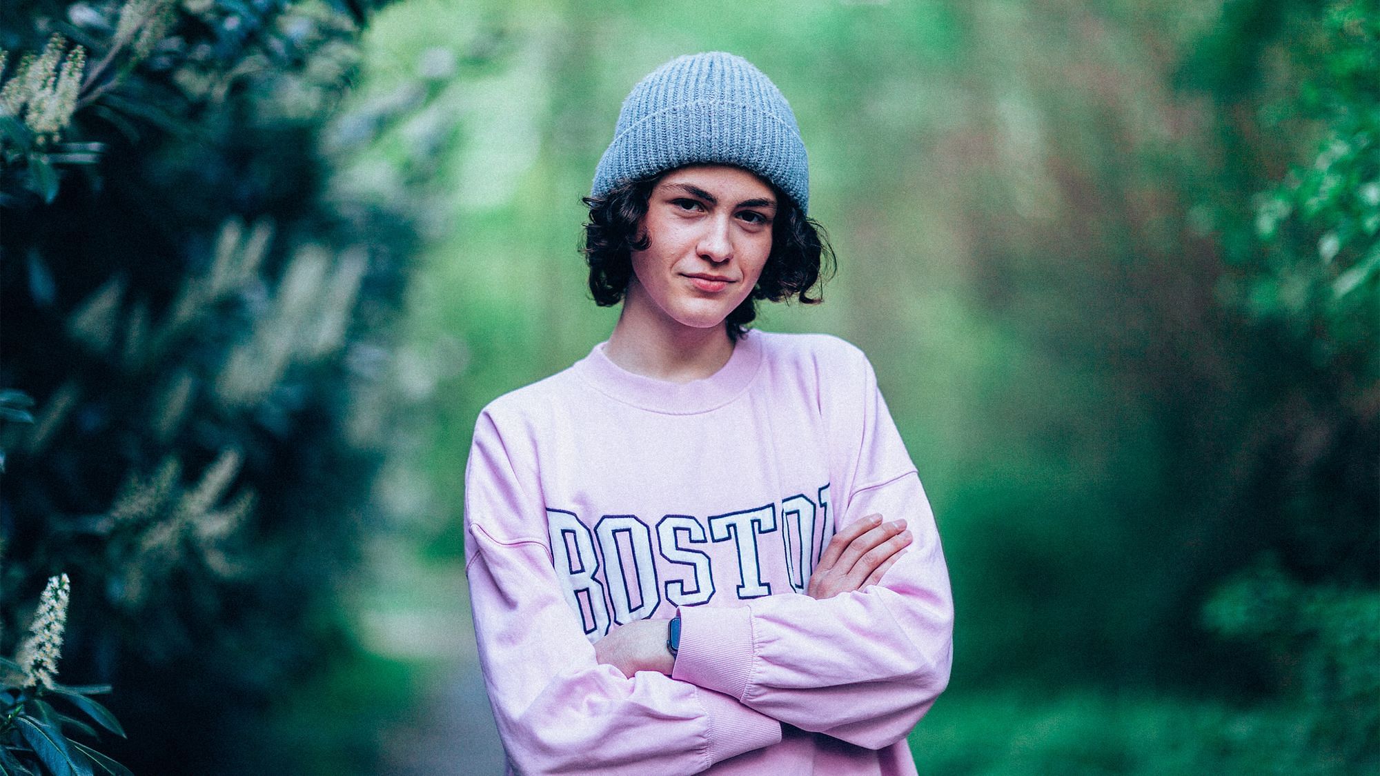 Ein Portrait von Gianni. Mit verschränkten Armen blickt er in die Kamera, er lächelt. Er trägt einen rosa Pulli und eine graue Mütze.