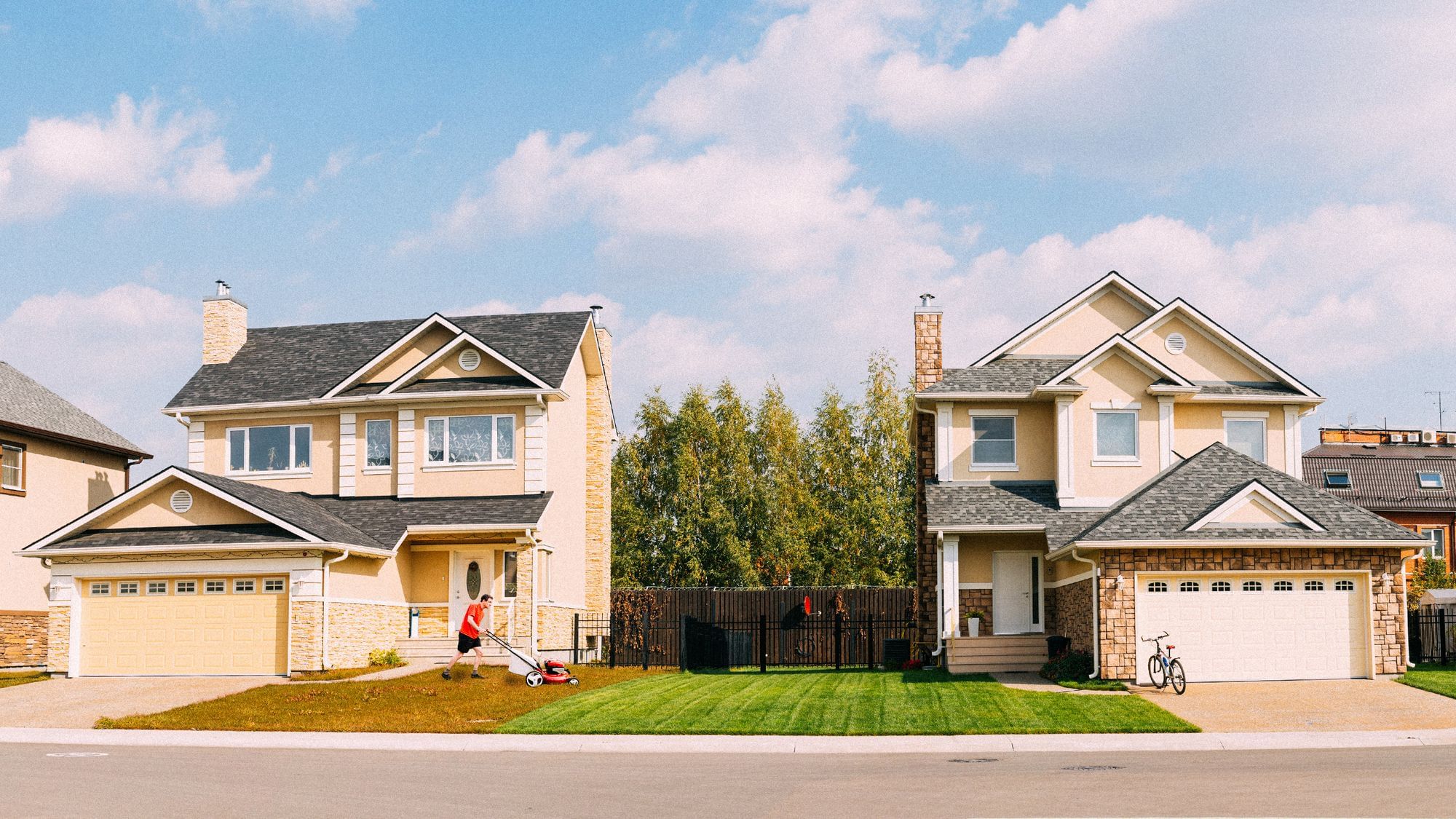 Zwei Häuser in der Vorstadt in Frontalansicht. Der Rasen links ist braun, der Rasen rechts ist grün. Der Rasen links wird gerade von einem Mann gemäht.