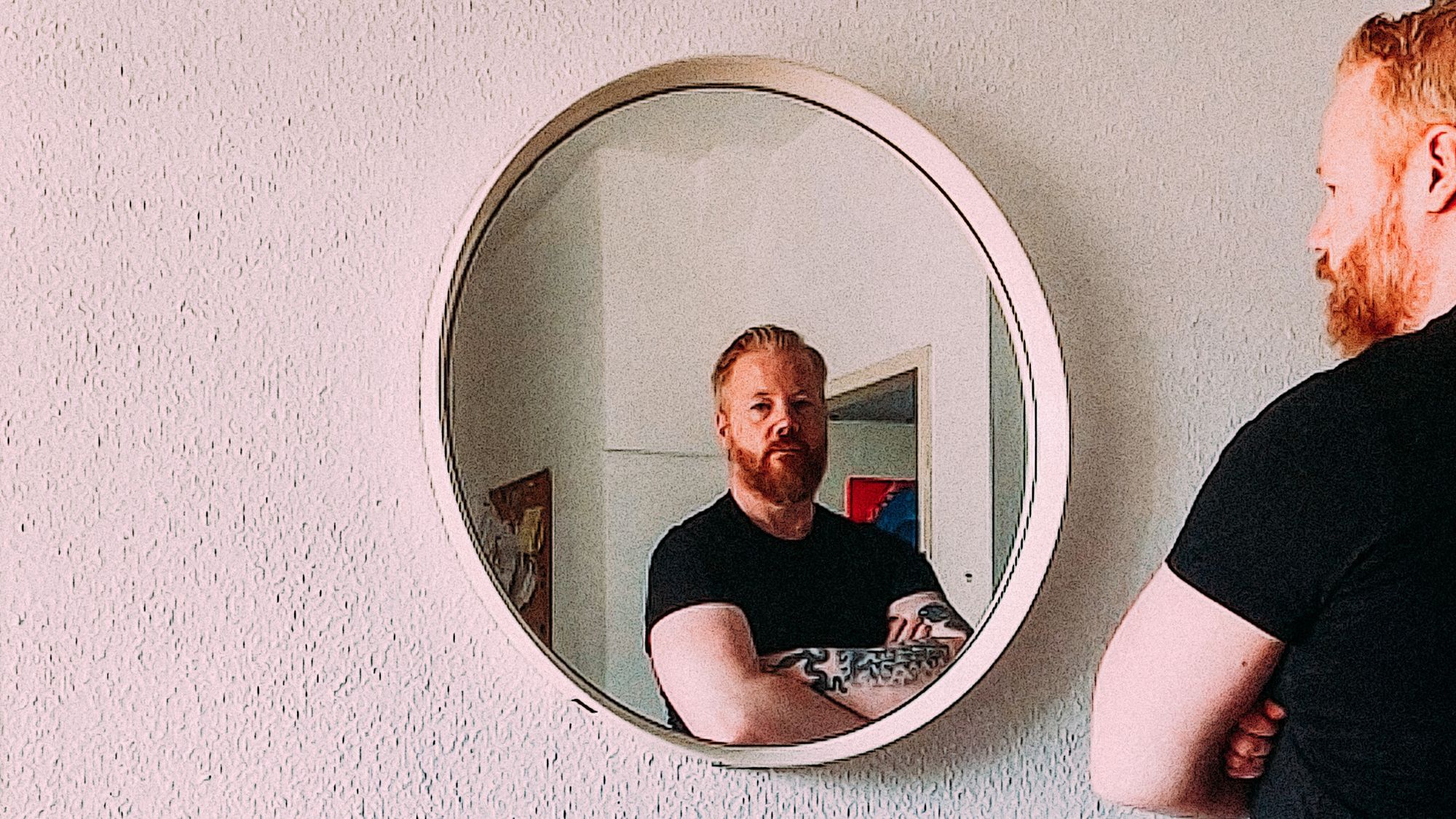 Martin steht mit verschränkten Armen vor einem Spiegel. Er blickt über den Spiegel in die Kamera.