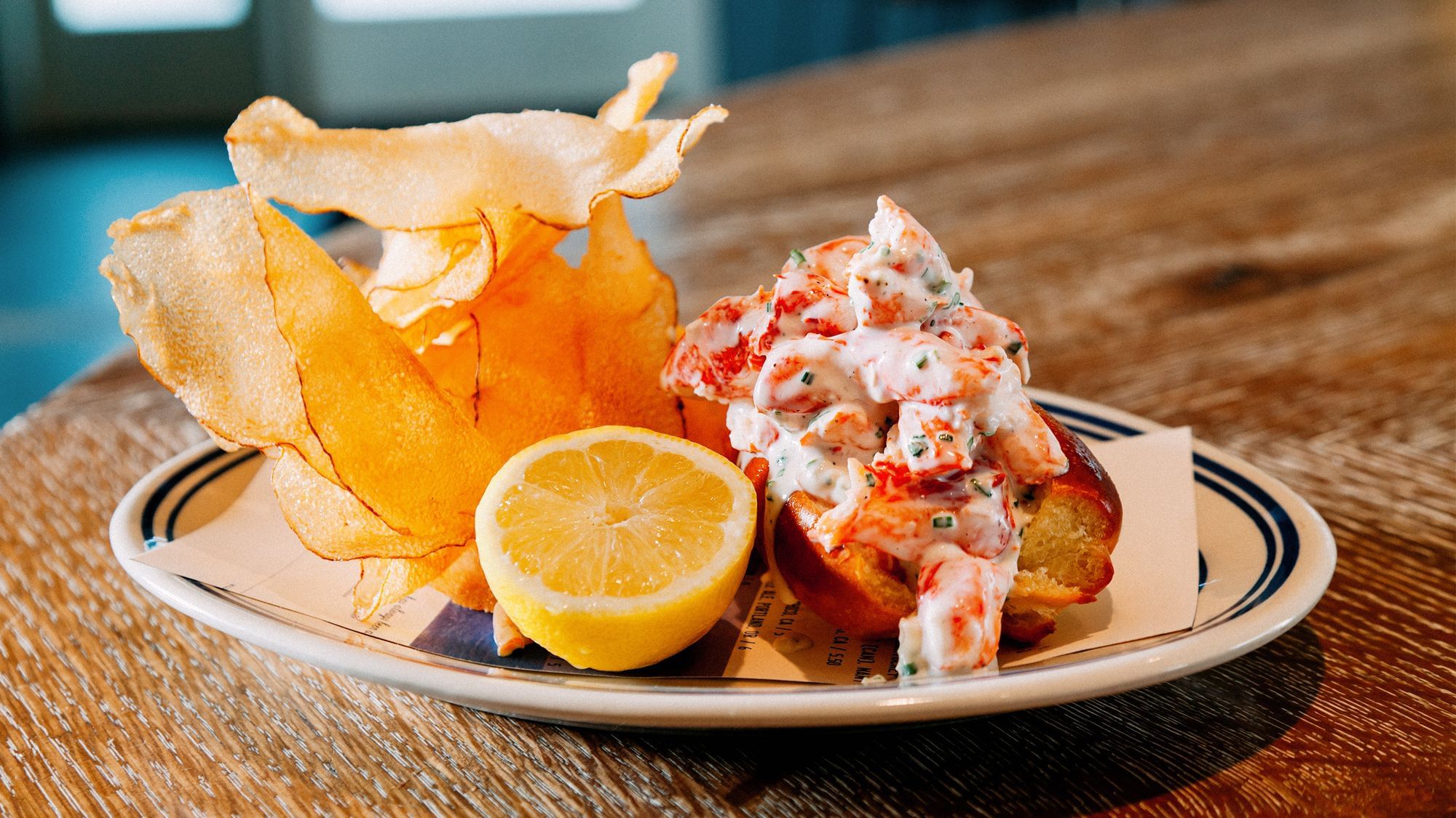 Man sieht ein Brötchen befüllt mit Hummerfleisch, daneben eine halb aufgeschnittene Zitrone und zu einem kleinen Turm drapierte Chips