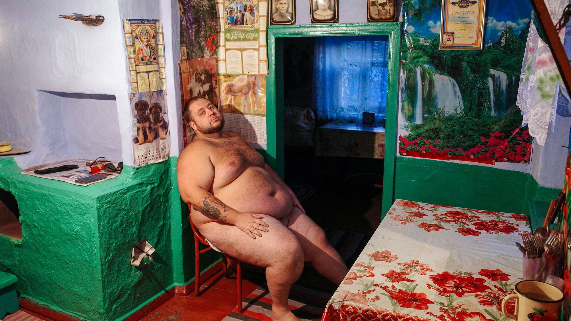 Wir sehen einen großen, dicken und nackten Mann, der auf einem Feld steht. Der Horizont brennt und der Mann ist ein rotes Licht getüncht.