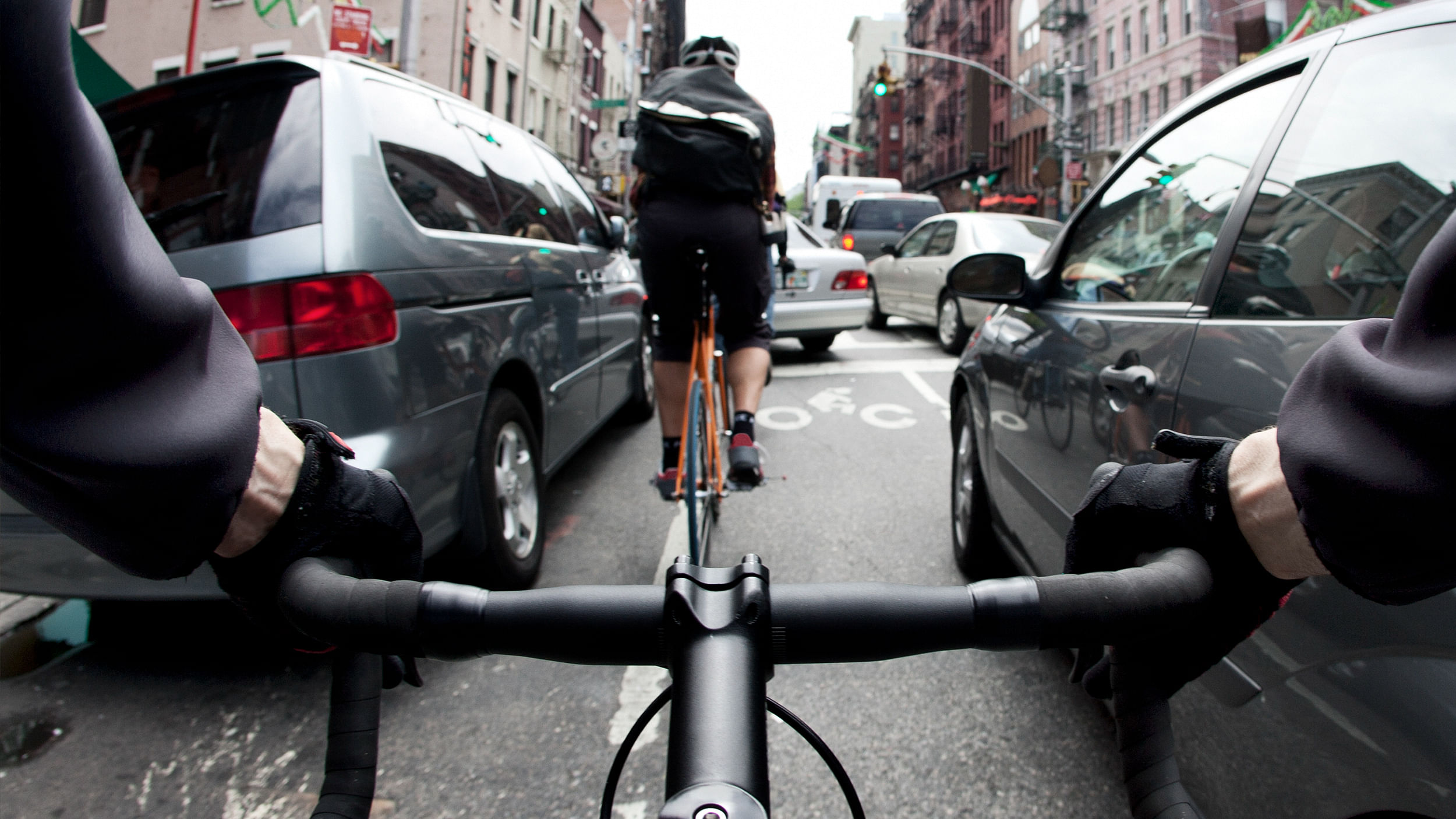 Ego-Perspektive eine Radfahrers. Man sieht seine Hände am Lenkrad. Vor ihm fährt ein weiterer Mensch auf der Straße, links und rechts ist nur wenig Platz bis zu den Autos. Im Hintergrund staut sich der Verkehr, für die Radfahrer gibt es kein weiterkommen.