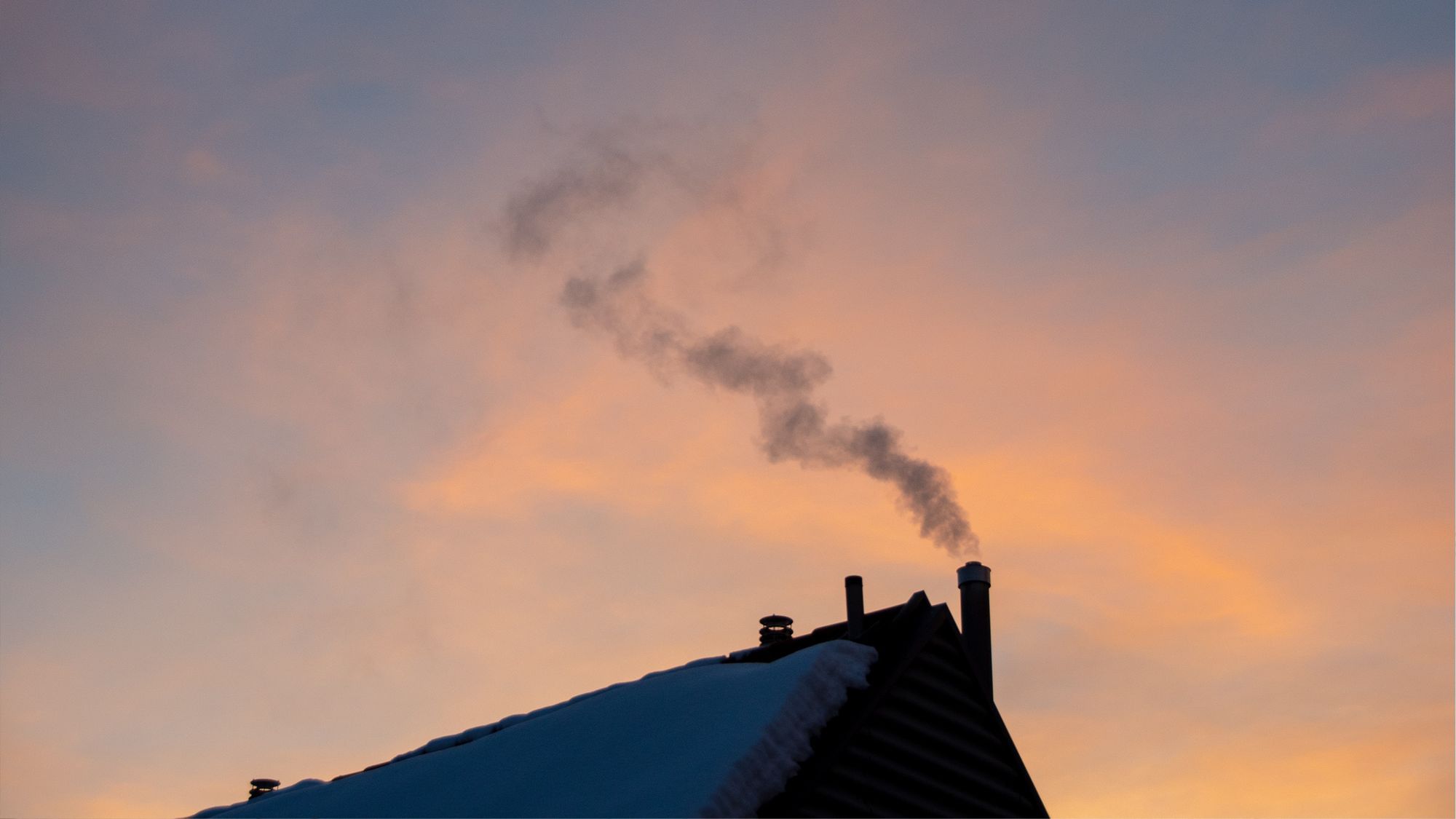 Ein schneebedecktes Dach bei Sonnenuntergang. Aus dem Schornstein steigt Rauch auf.