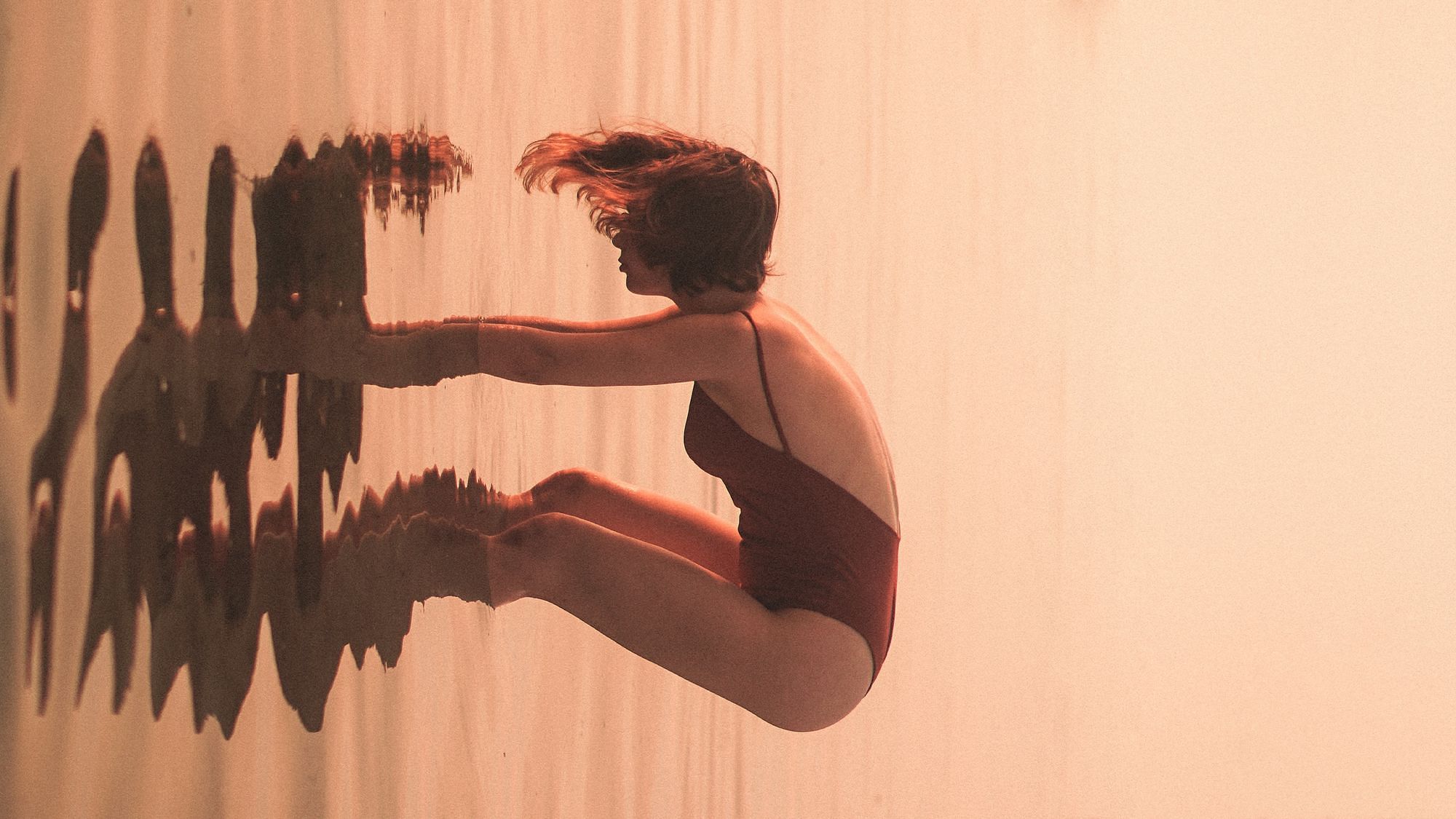 Eine Frau steht im Wasser und beugt sich hinunter, mit ihrem Händen ist sie im Wasser. Das Bild ist um 90 Grad gedreht, sodass der Wasserspiegel vertikal statt horizontal liegt.