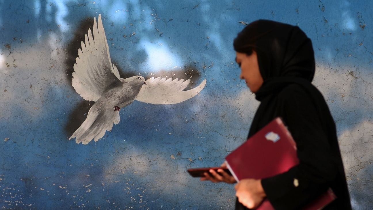 Eine Frau, die einen schwarzen Schal um den Kopf trägt, läuft eine Straße hinunter. Hinter ihr auf der Wand ist eine Friedenstaube vor blauem Hintergrund zu sehen.
