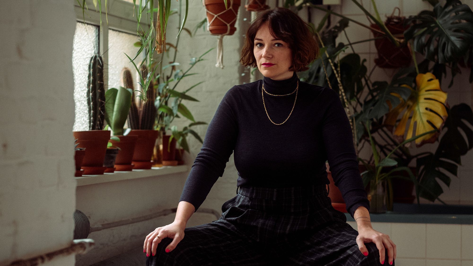 Mareice Kaiser trägt einen schwarzen Rollkragenpullover und eine schwarze Hose. Sie sitzt breitbeinig in einem Café, im Hintergrund ranken Pflanzen.