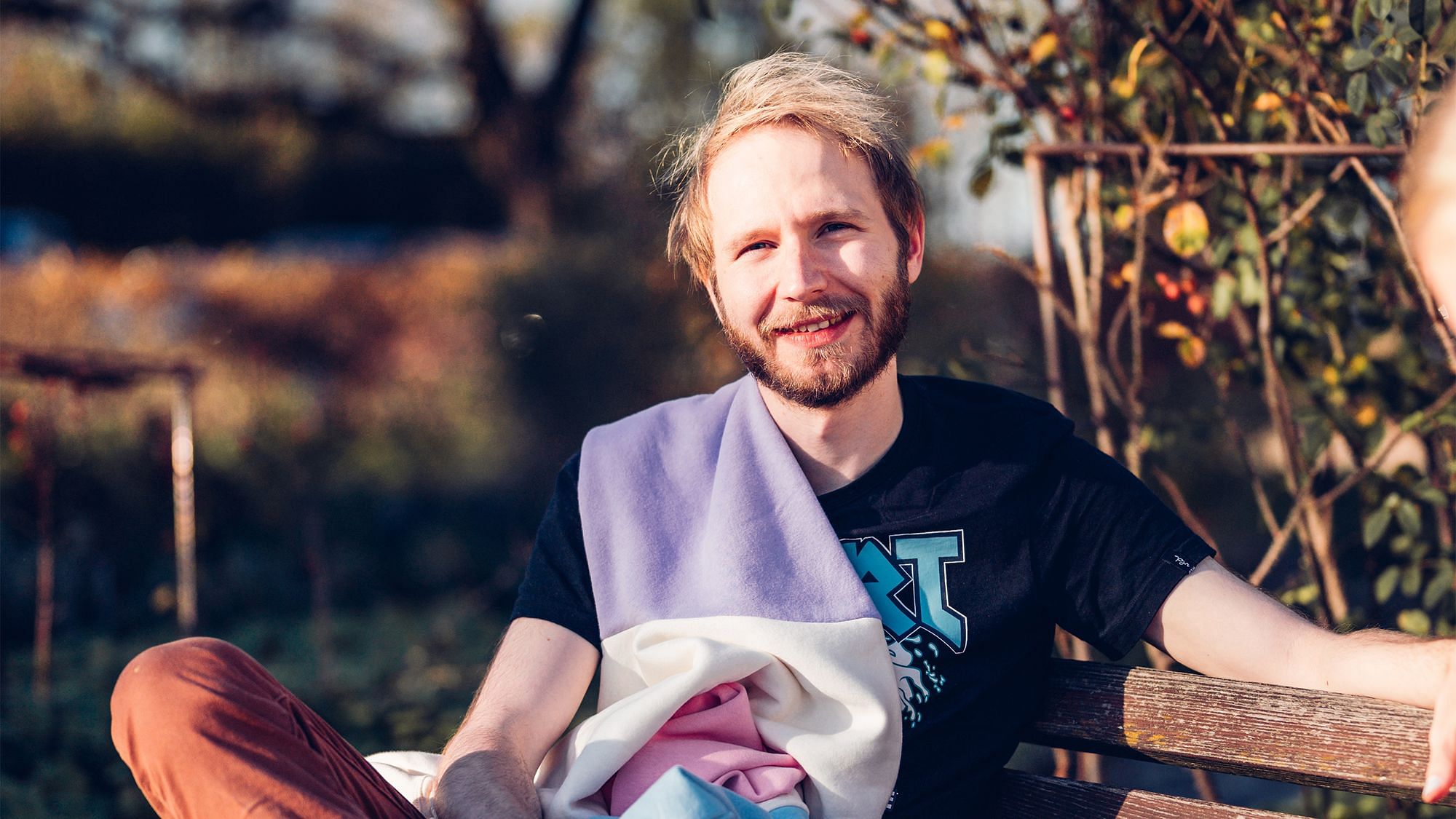 Eike sitzt auf einer Bank, er blickt in die Kamera. Er hat blond gefärbte Haare und trägt ein schwarzes Shirt, über seine Schulter hängt ein Bunter Pullover.