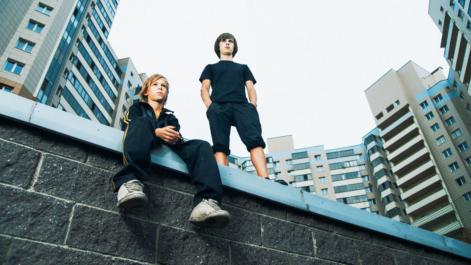 Zwei Jungs blicken in die Ferne. Einer sitzt auf einer Mauer, der andere steht neben ihm. Sie befinden sich in einer Plattenbausiedlung.