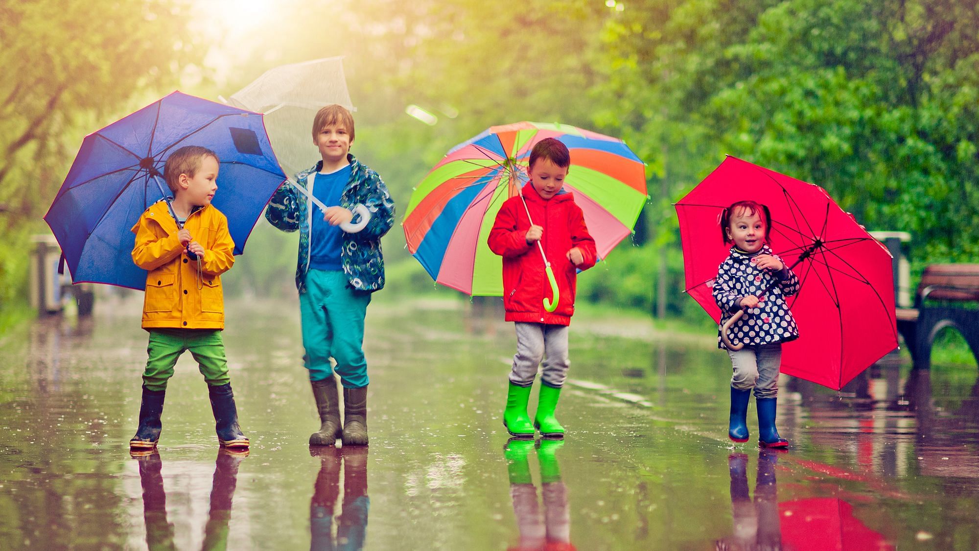 Vier Kinder stehen im Regen. Sie tragen bunte Regenkleidung und Regenschirme.