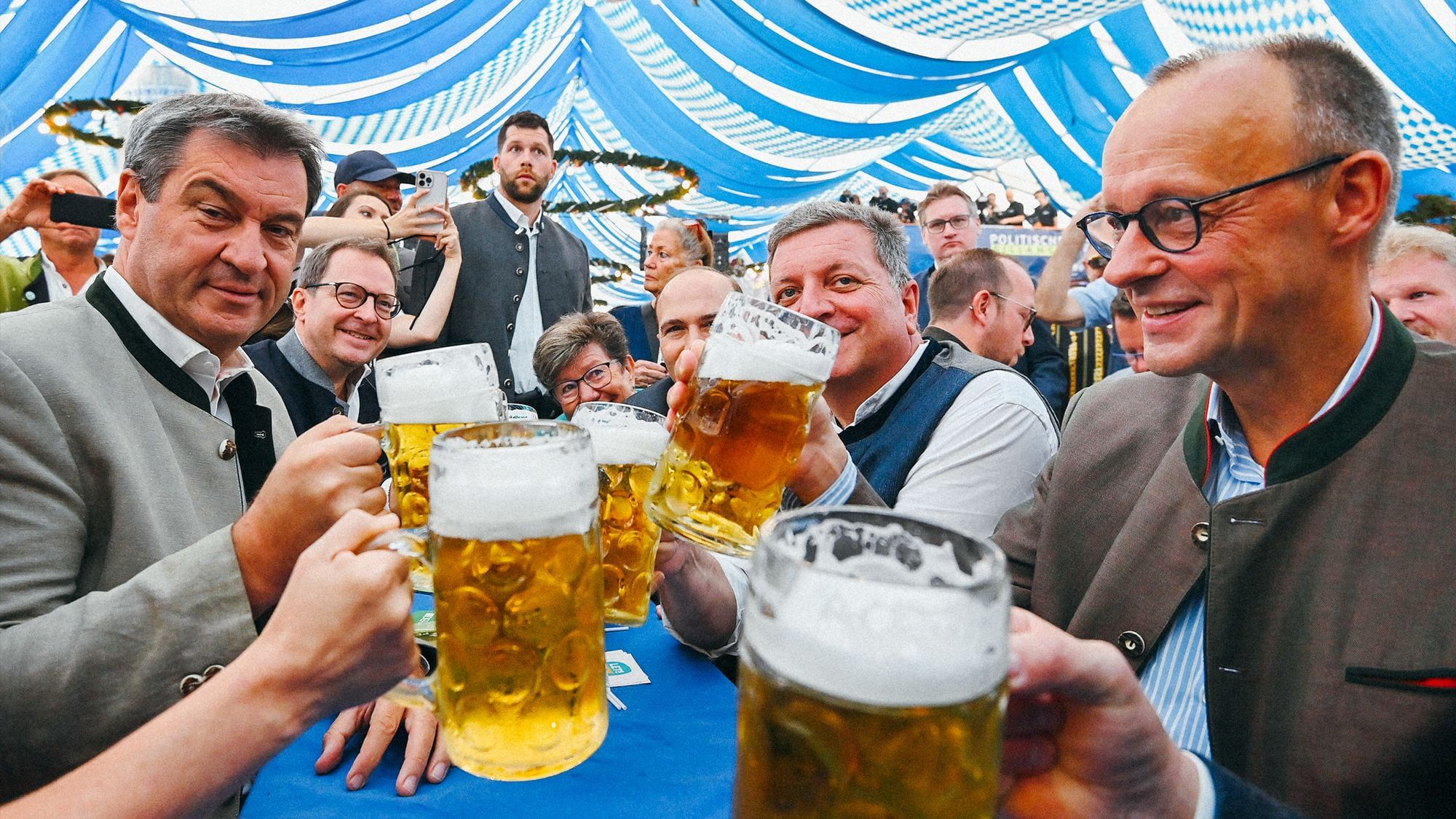 Markus Söder und Friedrich Merz prosten sich mit einer Maß Bier in der Hand in einem bayerischen Bierzelt zu 