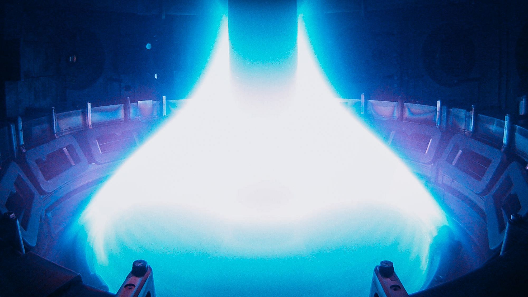 Foto vom Inneren eines Kernfusionsreaktors. Man sieht blau leuchtendes Plasma, das sich kegelförmig um eine senkrechte Metallstange bildet.