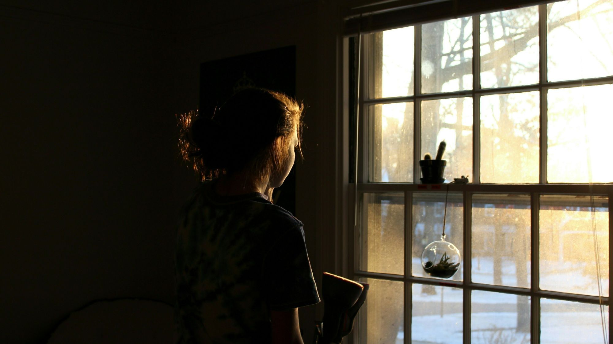 Frau steht allein in einem dunklen Zimmer und schaut aus dem Fenster in einen Garten