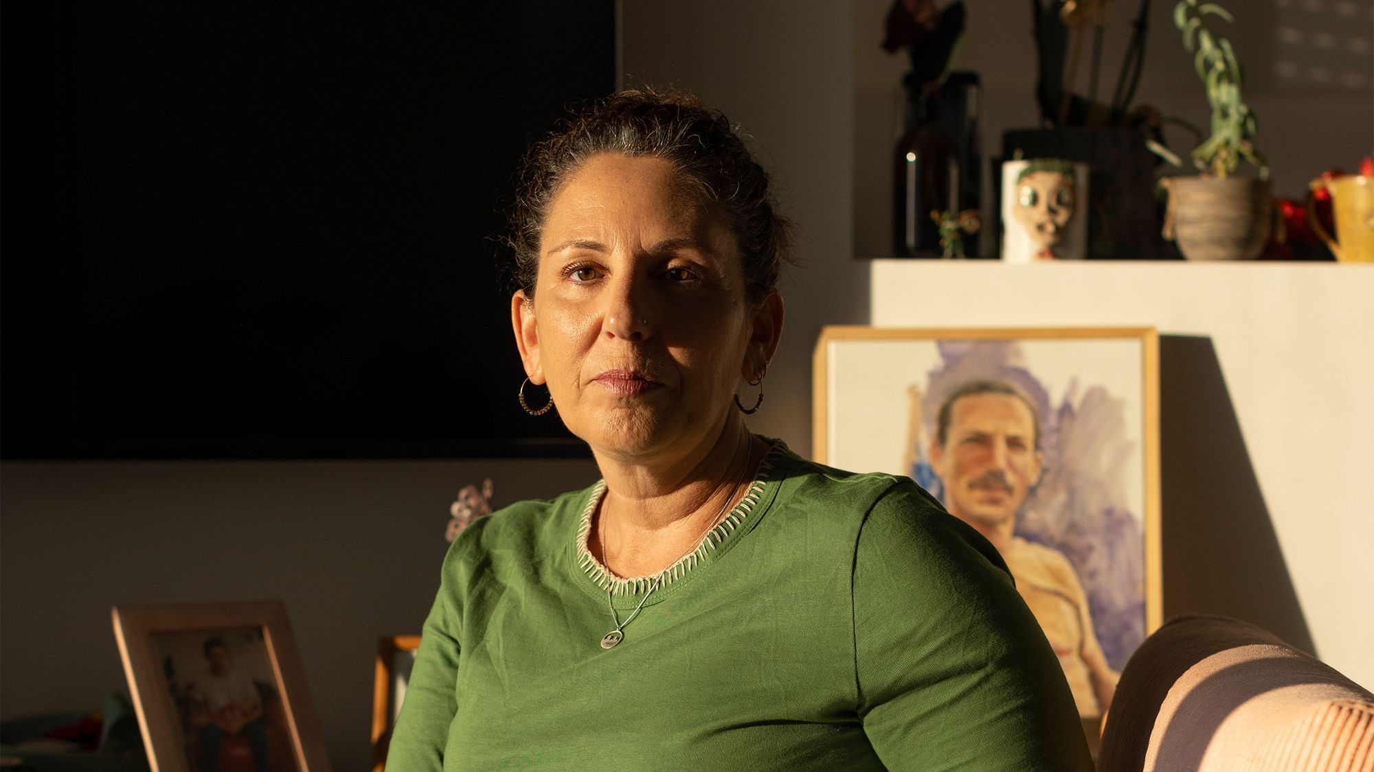 Eine Frau blickt in die Kamera. Sie trägt ein grünes Oberteil. Im Hintergrund stehen Bilder.