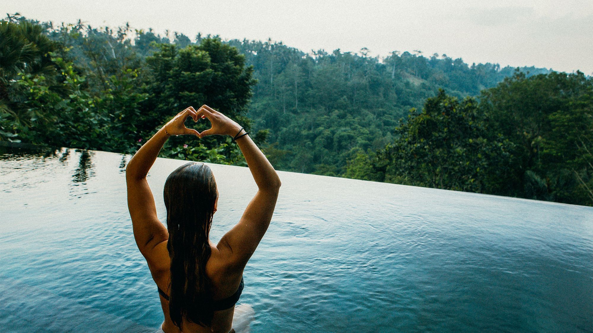 Eine Frau sitzt in einem Infinity Pool, von dem aus man auf einen Wald blicken kann. Sie hält die Arme über den Kopf und formt ihre Hände zu einem Herzen.