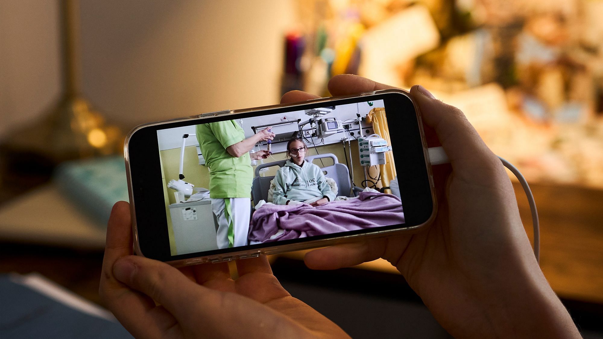 Eine Hand hält ein Handy in die Kamera. Darauf ist ein Mädchen zu erkennen, das in einem Krankenhausbett liegt. Es ist umgeben von Maschinen, eine Pflegeperson steht daneben.