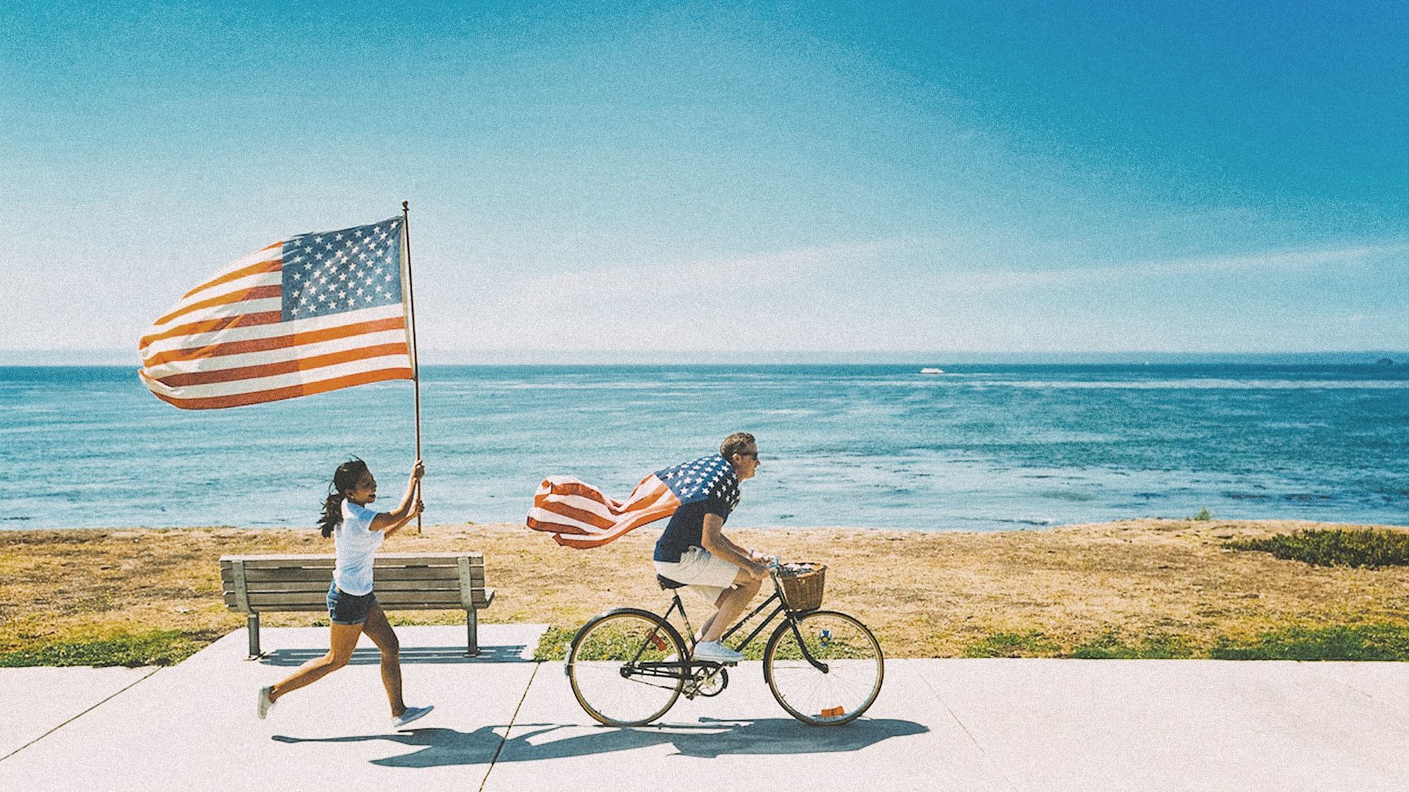 Zwei Menschen auf einer Straße am Meer. Ein Mann fährt Fahrrad und hat eine amerikanische Flagge umgebunden, eine Frau läuft im hinterher während sie eine amerikanische Flagge in die Höhe hält. 