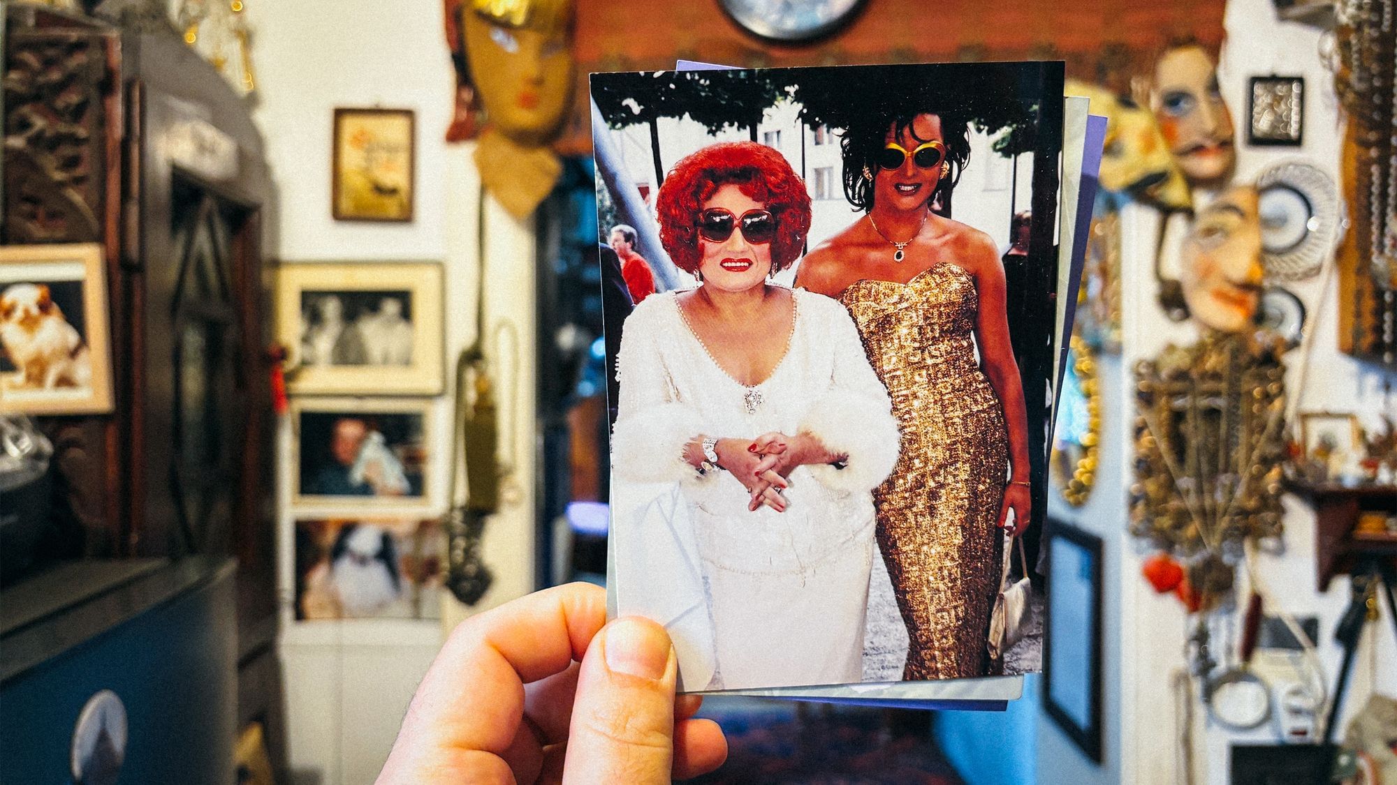 Eine Hand hält ein Foto von zwei Travestie-Künstlerinnen in die Kamera. Eine ist in einem weißen Kleid und roter Perücke gekleidet, die andere in einem goldenen Kleid. Im Hintergrund sieht man ein vollgestopftes Zimmer.
