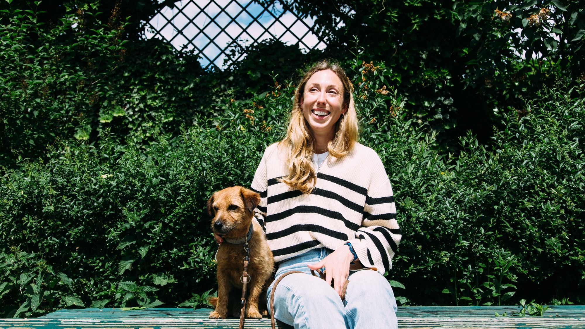 Nadine sitzt neben ihrem Hund, ein Terrier-Mix, auf der Bank. Sie lächelt und trägt Pullover mit Schwarz-Weißen Streifen.