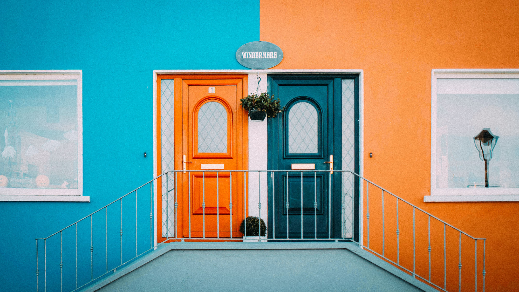 Eine mittig geteilte Hausfront mit zwei Türen. Eine Seite ist blau mit orangener Tür, die andere orange mit blauer Tür.