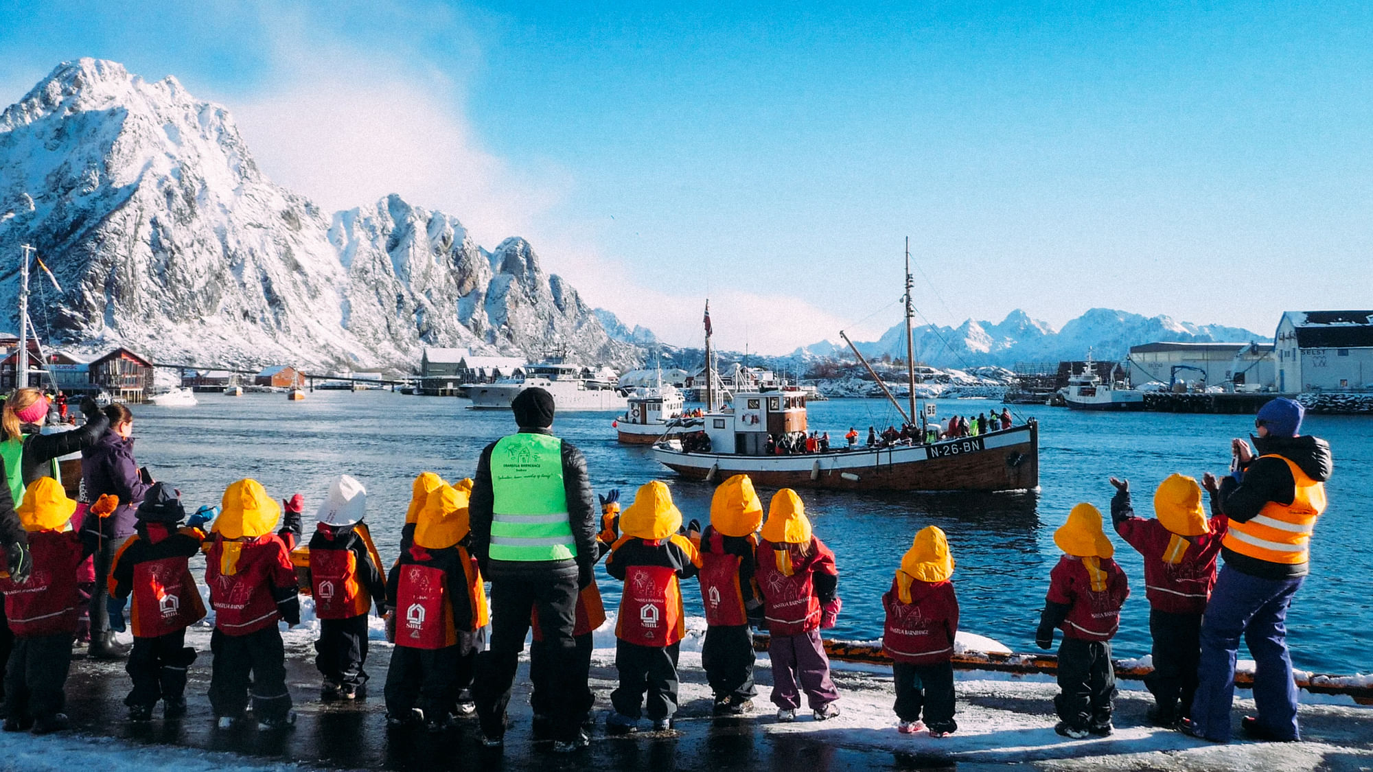Eine Gruppe Kindergartenkinder mit Betreuer:innen steht an einem Hafen und schaut auf die Schiffe. Im Hintergrund ist ein Berg zu sehen.