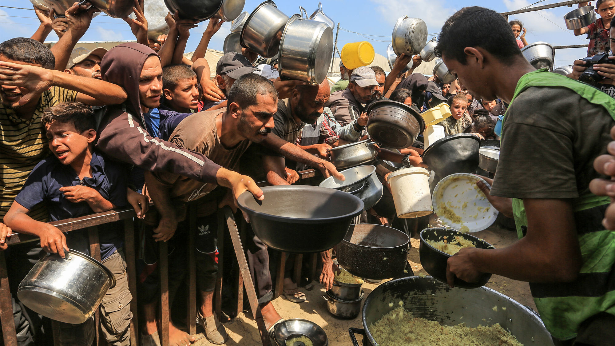 Ein Hilfsarbeiter verteilt gekochten Reis an Menschen. Sie stehen in einer Menge hinter einem Zaun und halten Töpfe in der Hand.