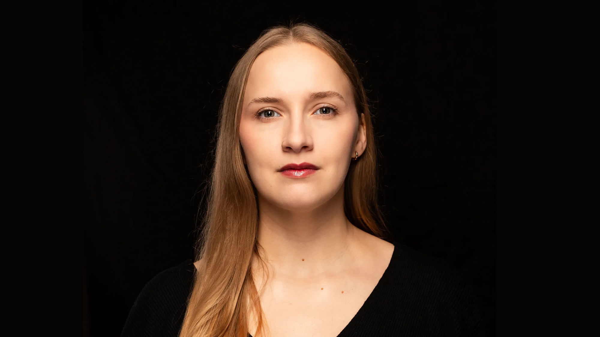 Eine Frau mit braunen Haaren blickt in die Kamera. Sie ist schwarz gekleidet. Auch der Hiinterrund ist schwarz.