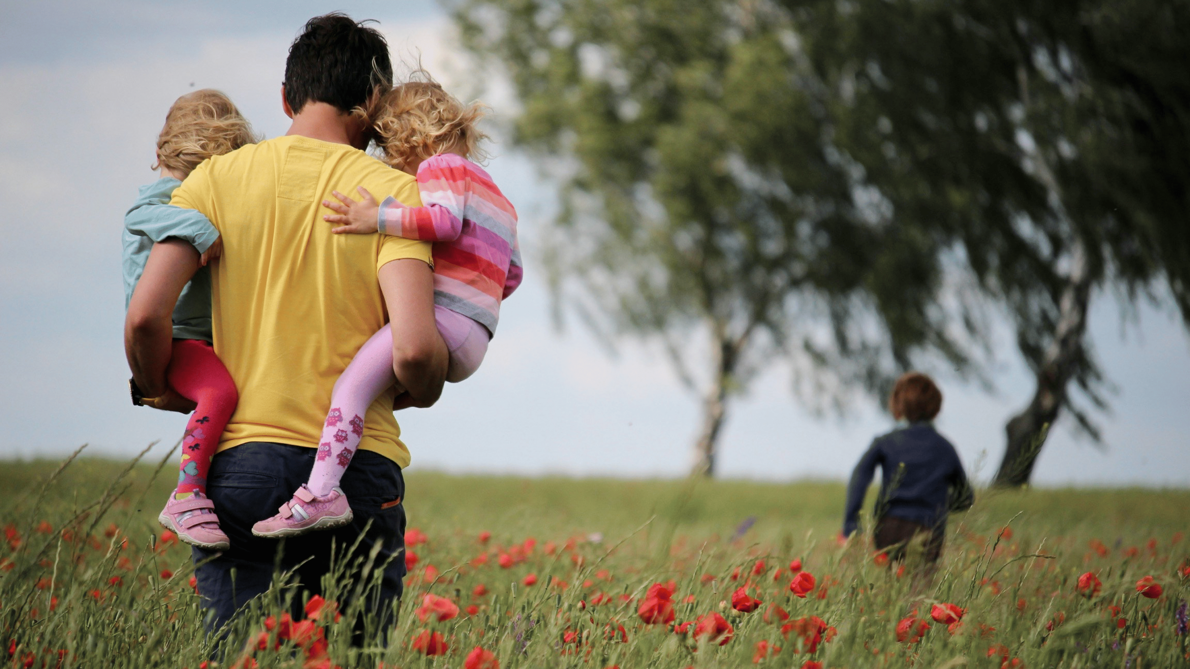 Mann mit zwei Kindern auf dem Arm, ein weiteres Kind rennt richtung Baum auf der rechten Seite